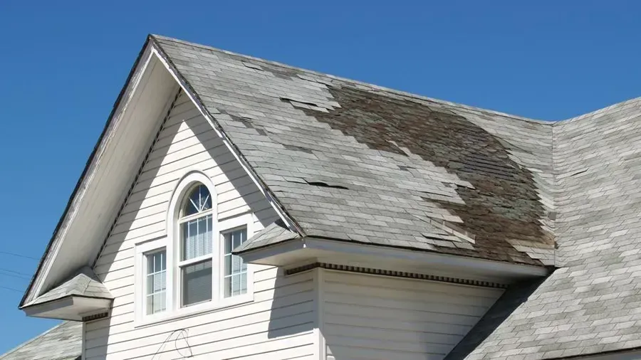 Damaged asphalt shingle roof on a white house with a gabled roof and arched window.