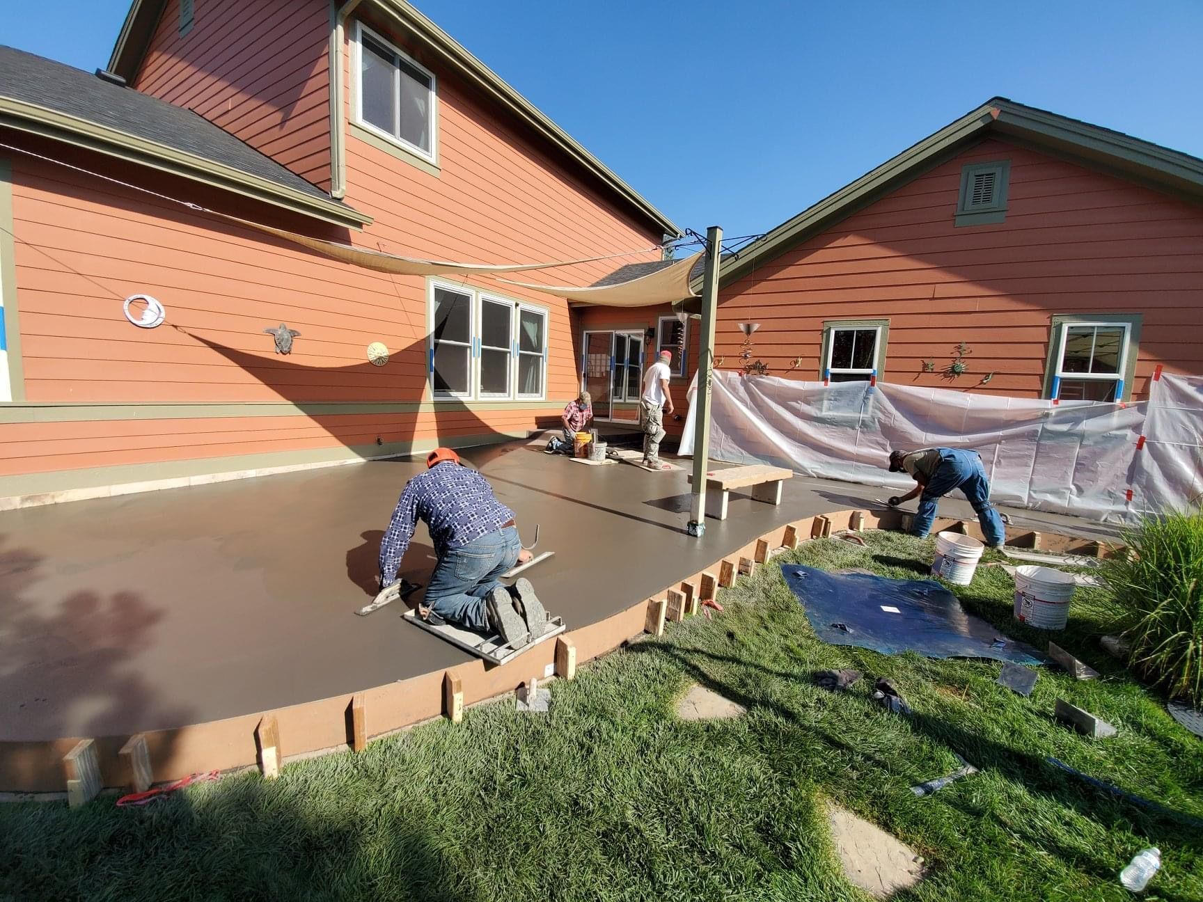 Two men are working on a concrete deck in front of a house.