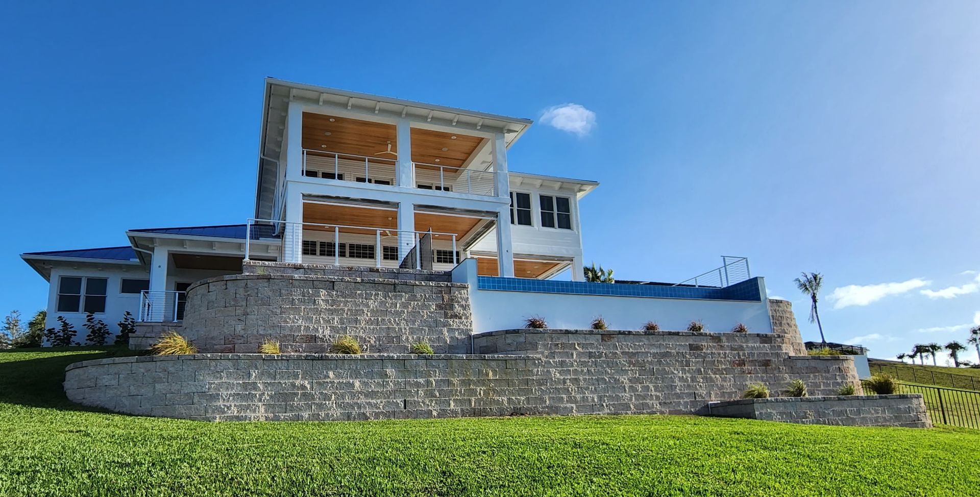 Two-story white house with stone retaining wall, a pool, and blue sky on a sunny day.