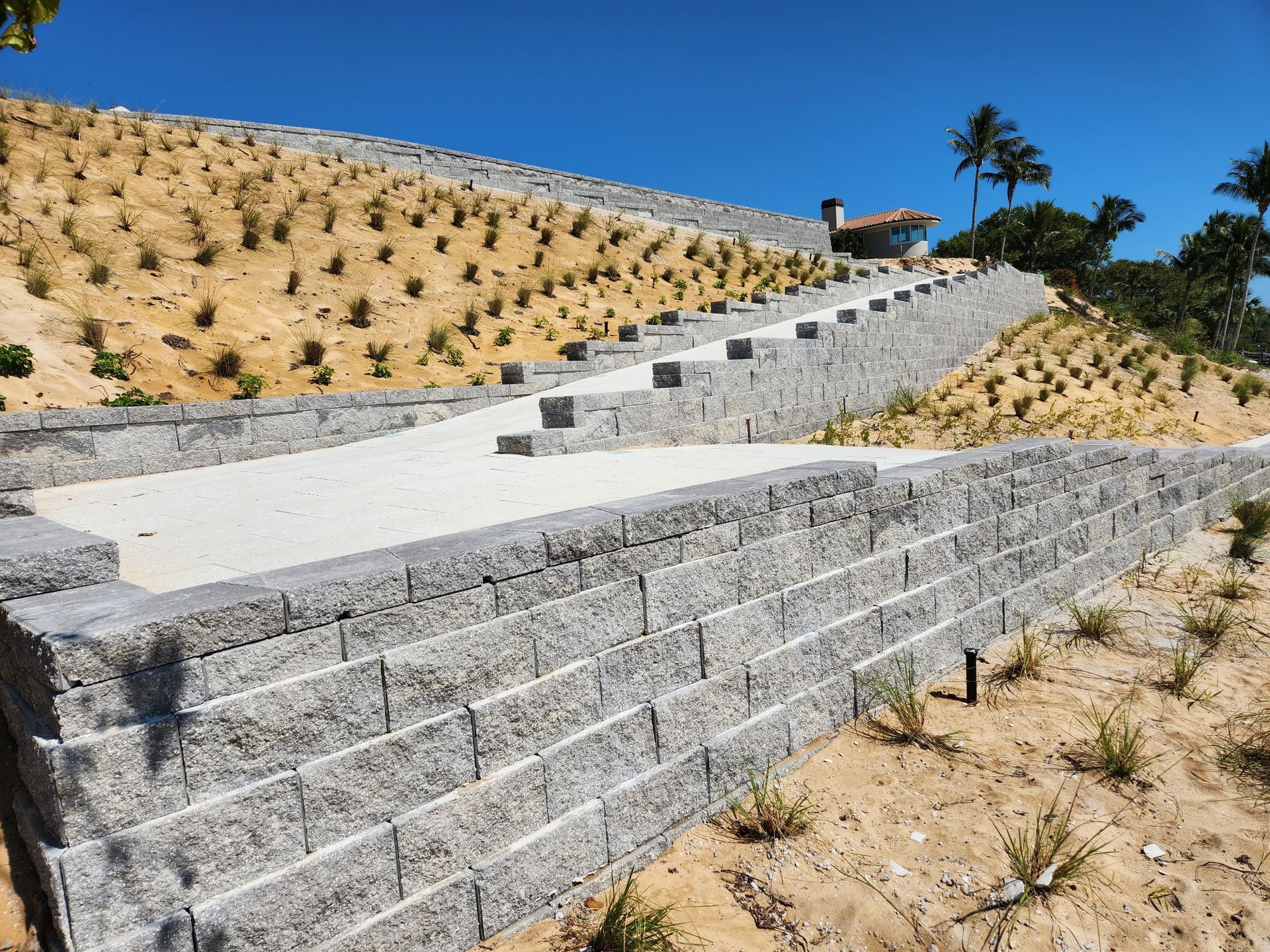 Stone retaining walls with a concrete pathway ascending a hillside, under a blue sky.