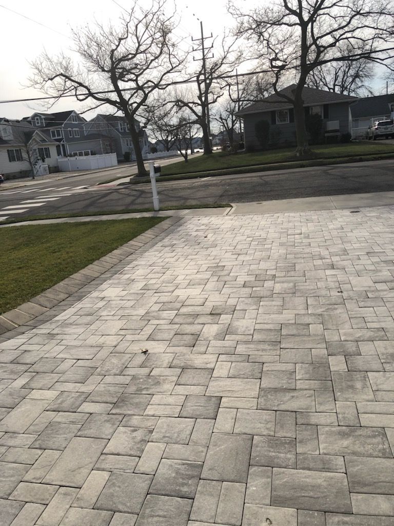 Brick paver driveway leading to a suburban street with houses and bare trees.