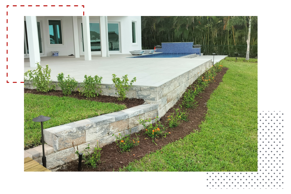 A modern home with a stone retaining wall borders a patio with landscaping; green grass.
