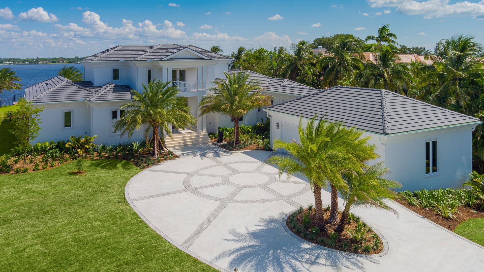 White waterfront mansion with palm trees, circular driveway, and water view.