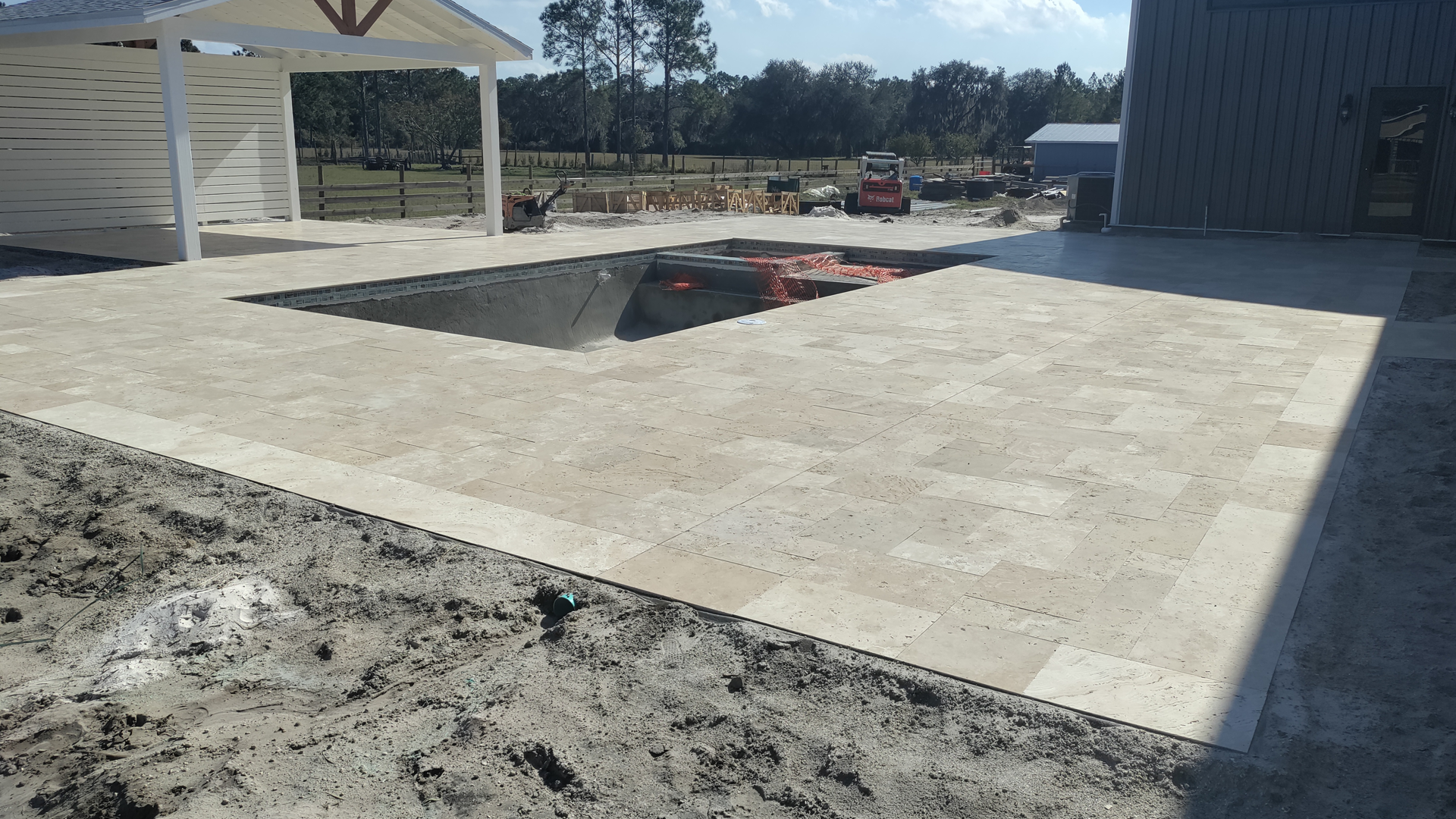 A light stone patio surrounds a dark pool.  Construction site with a white structure and gray building in the background.