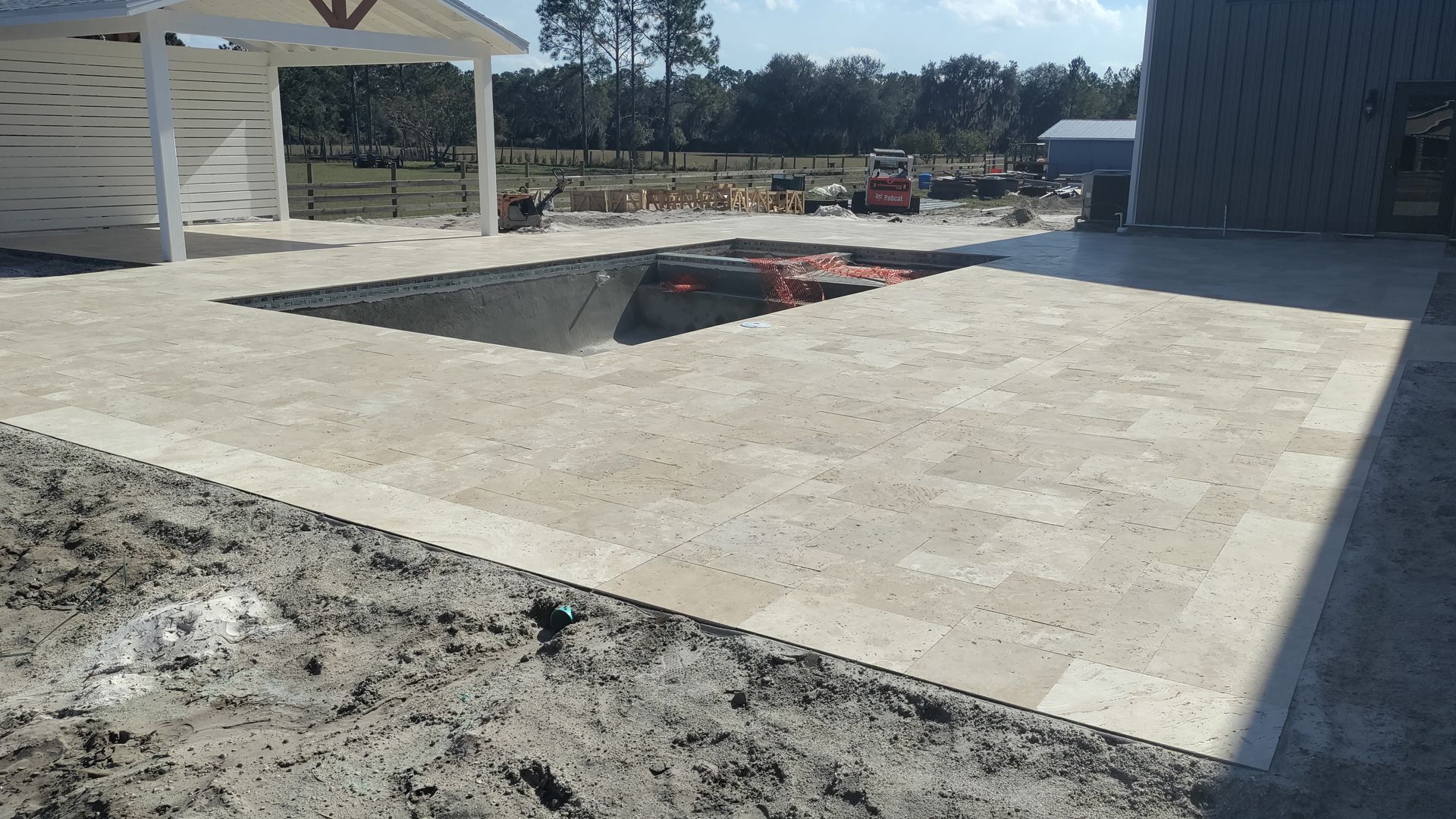 Paver patio with a square cut-out for a pool, surrounded by dirt; a white structure is in the background.