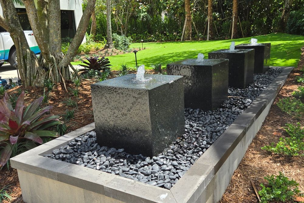 Four dark stone cube fountains with water spouts in a landscaped garden bed.
