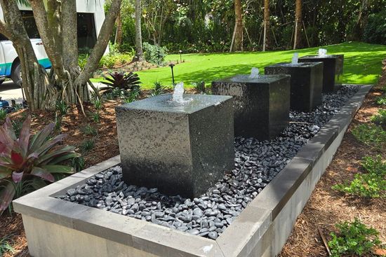 Four dark stone cube fountains with water spouts in a landscaped garden bed.