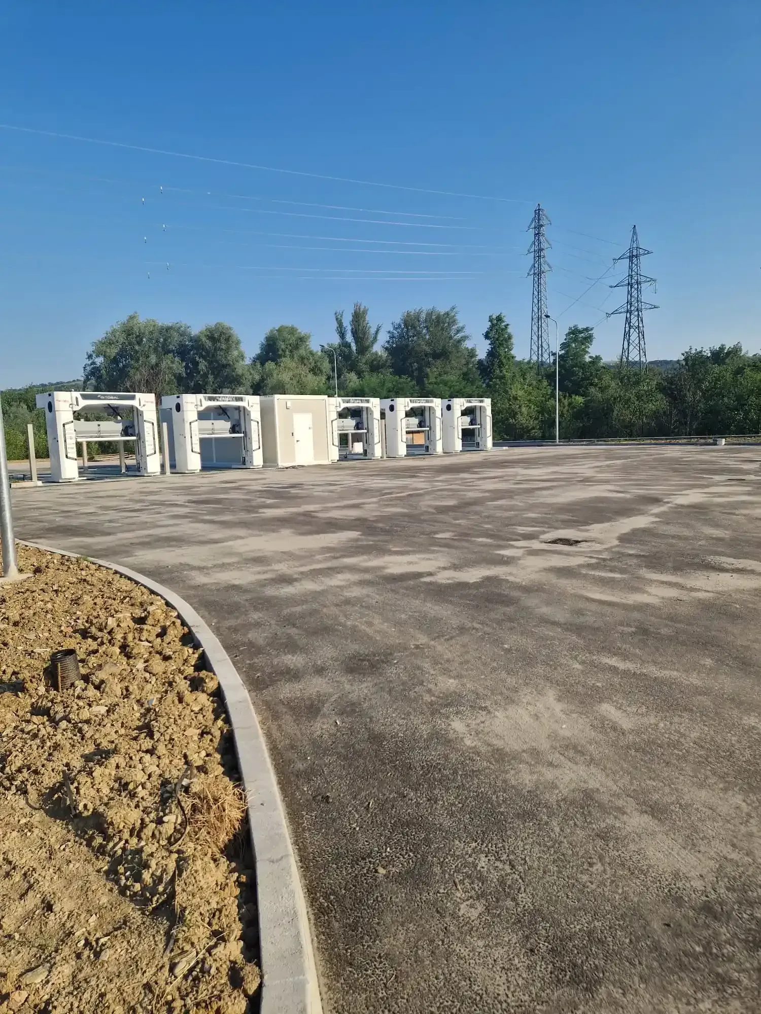 Una fila di stazioni di ricarica elettriche bianche sull'asfalto, con linee elettriche e alberi sullo sfondo sotto un cielo azzurro.