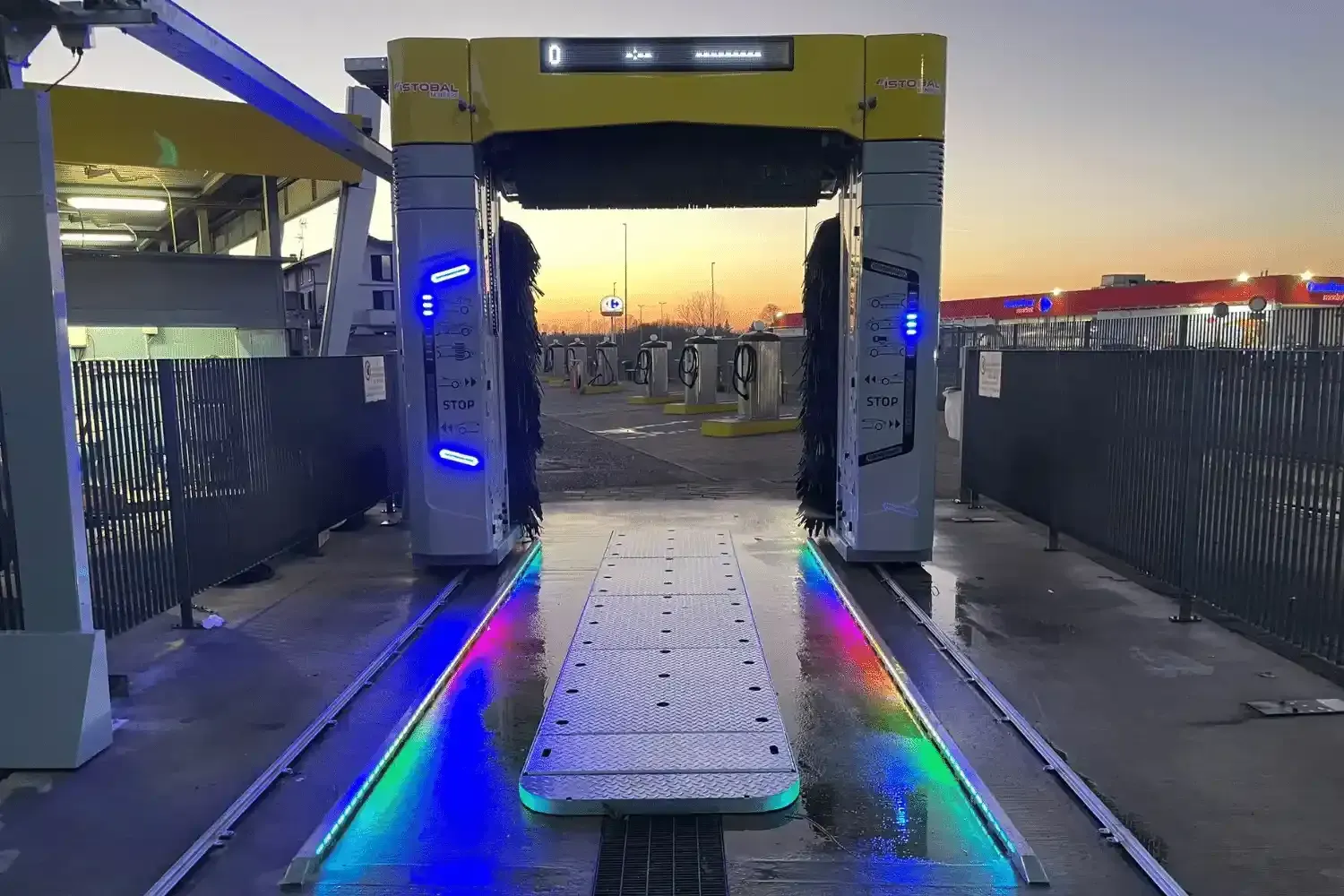 Autolavaggio, vista dell'ingresso con luci colorate e spazzole, ambientato all'aperto con cielo al tramonto.