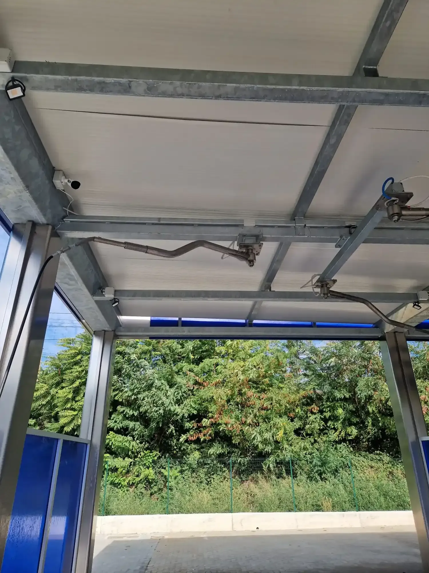 Area di lavaggio auto con tetto in metallo, pannelli laterali blu e vegetazione visibile sullo sfondo.