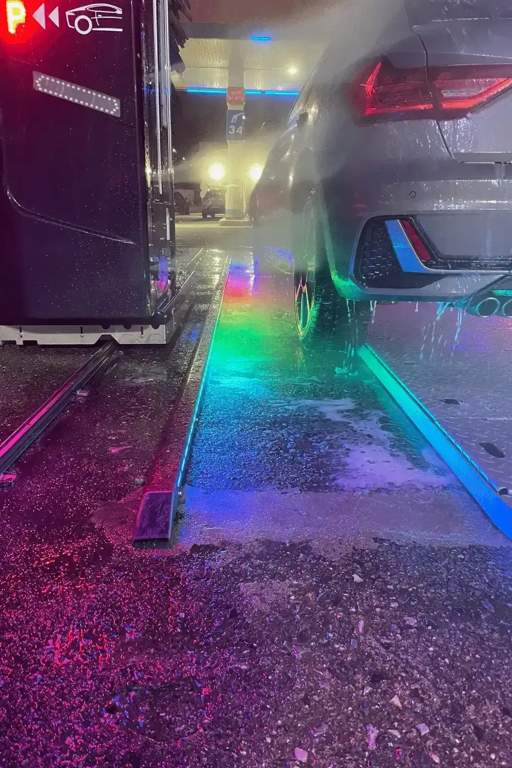 Auto in fase di lavaggio in un autolavaggio, illuminata da luci colorate sul pavimento.