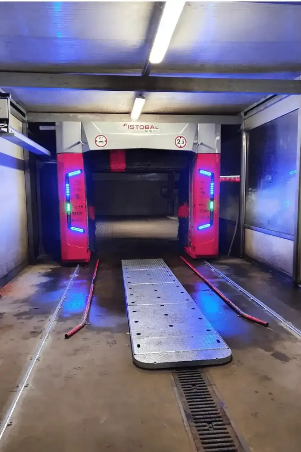 Area di lavaggio auto con macchinari rossi e grigi, luci blu e una piattaforma.