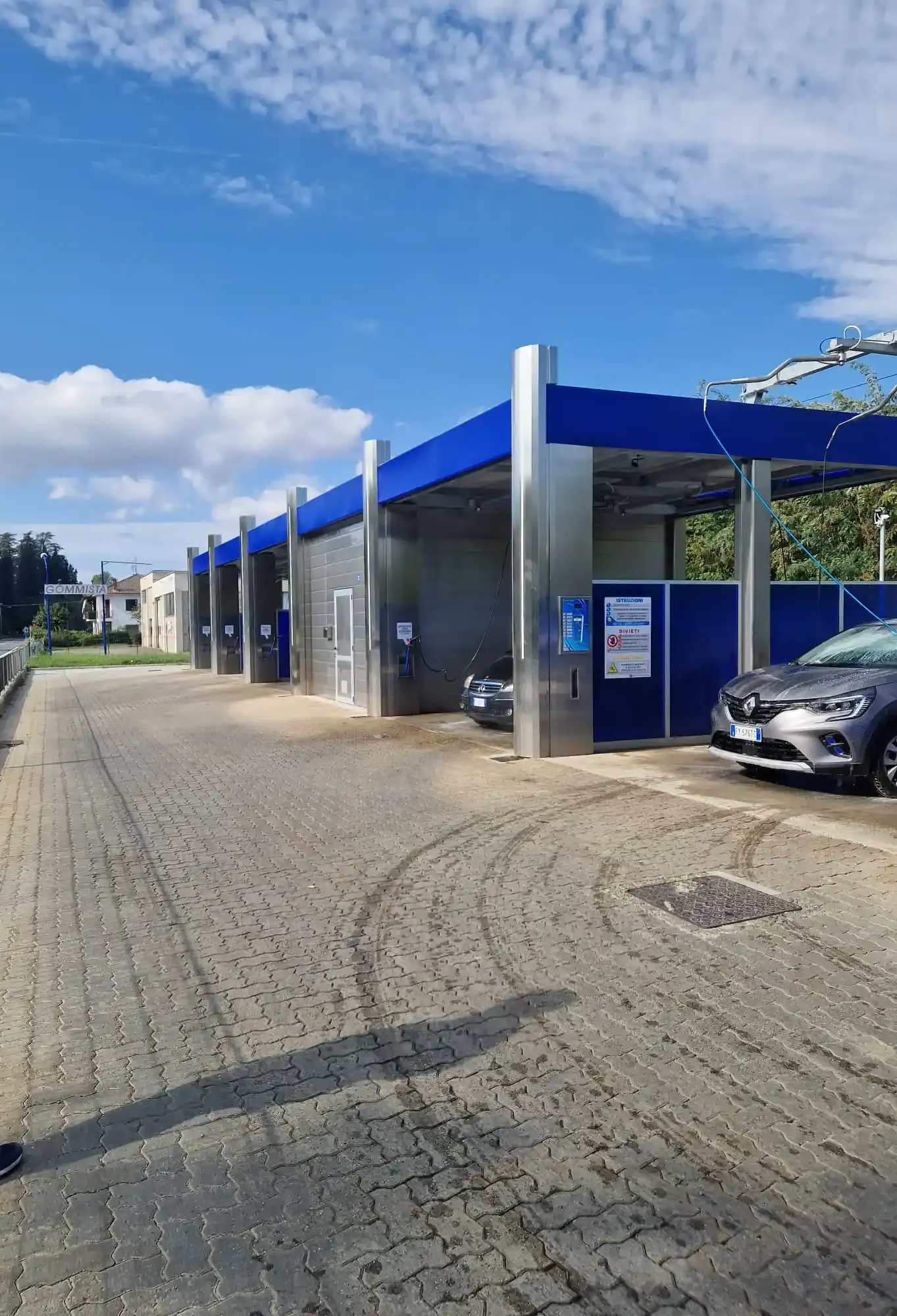 Autolavaggio con più postazioni, pavimento in mattoni grigi, strutture blu e argento sotto un cielo azzurro.