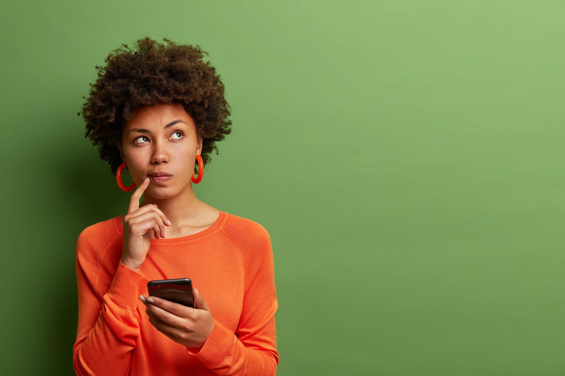 a woman in an orange sweater is holding a cell phone and looking up .
