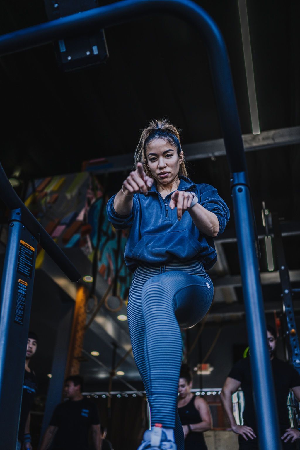 Woman points towards the viewer while on a curved treadmill. Wearing athletic wear in a gym setting.