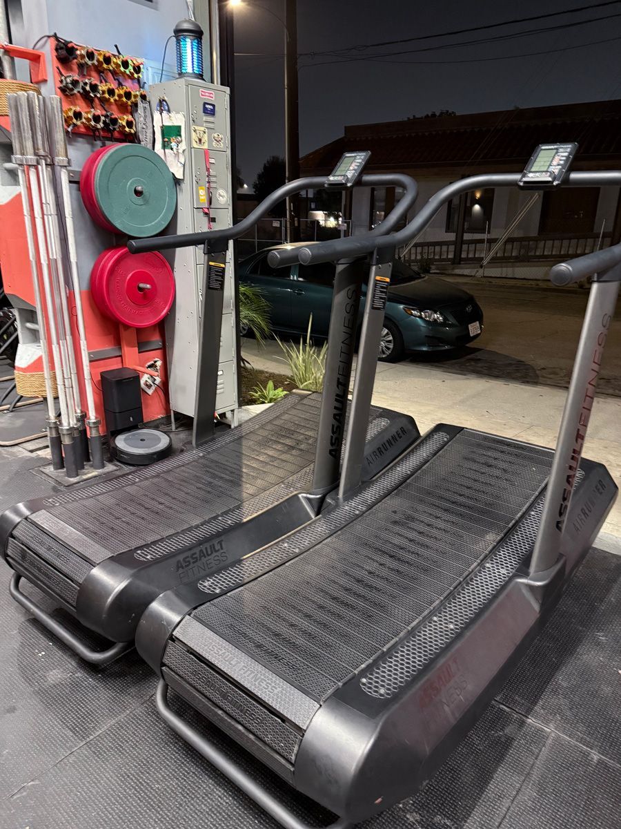 Two curved treadmills side-by-side in a gym. Grey machines with black treads, weights and equipment in background.