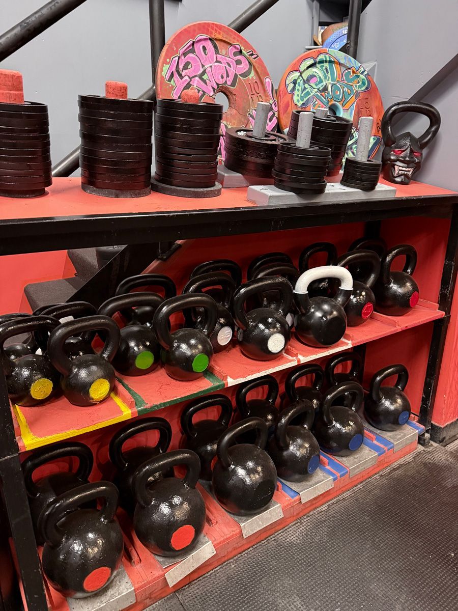 Shelves of black kettlebells with colored dots and weight plates in a gym.