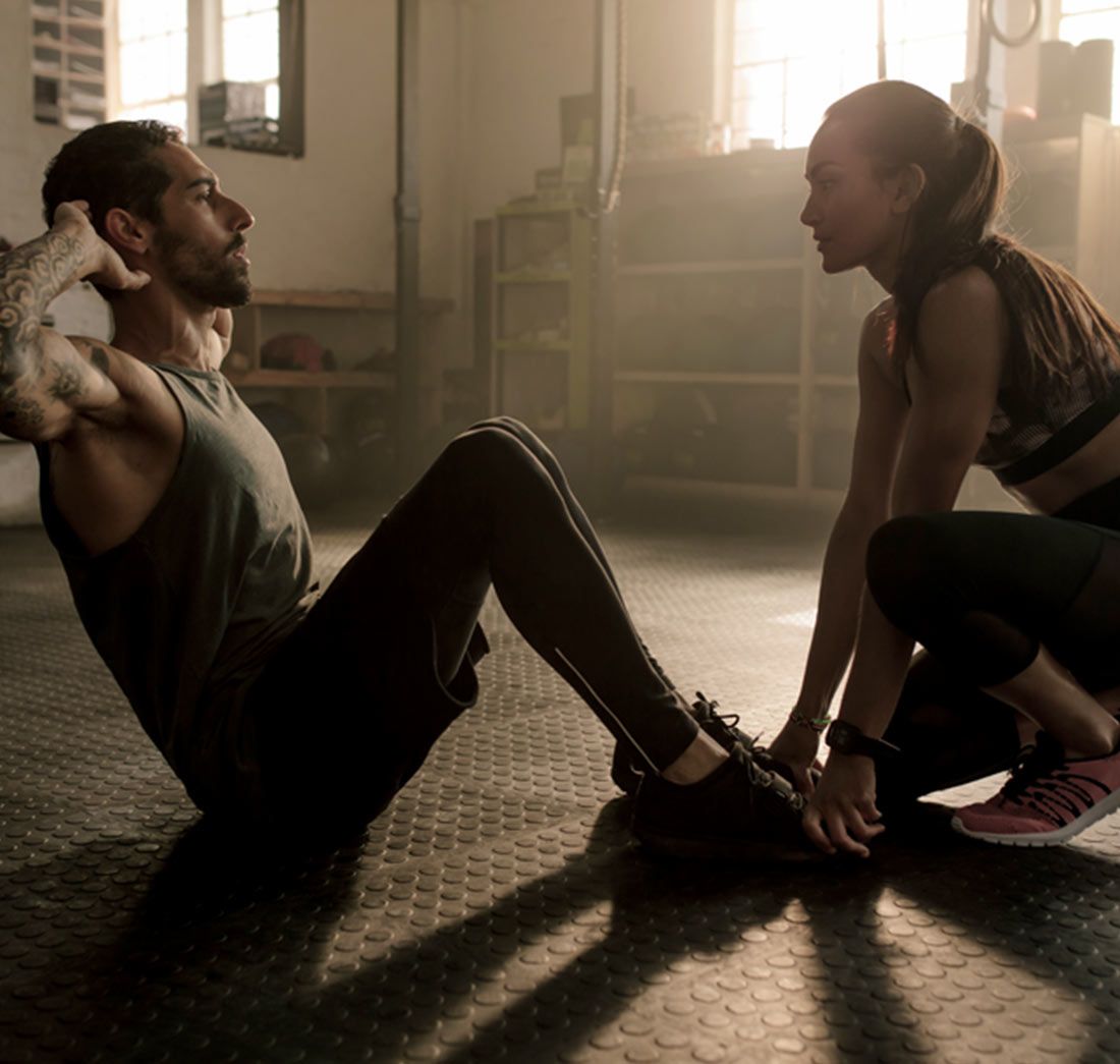 Man doing sit-ups with assistance from a trainer in a gym, working out on a black mat.
