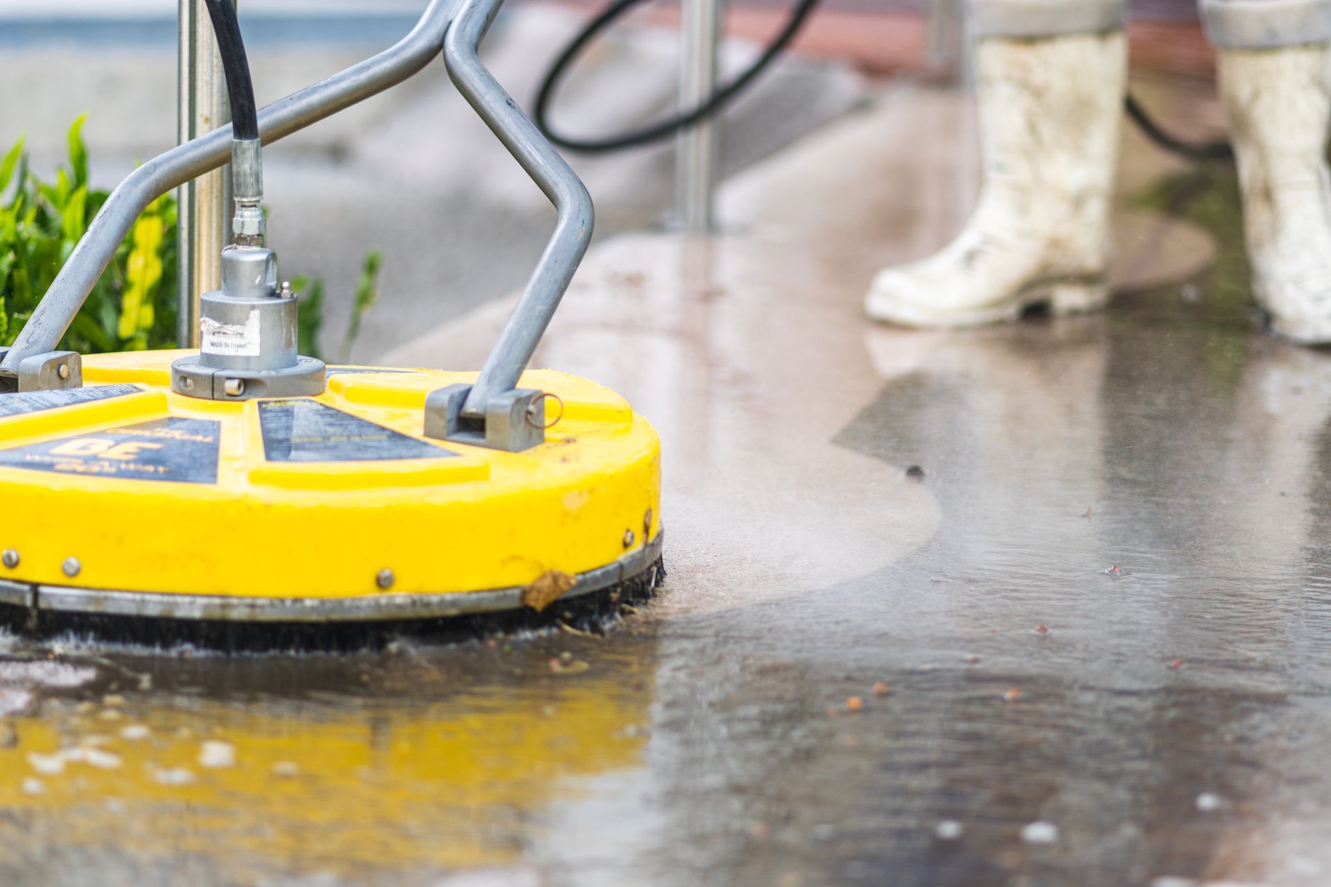 A person in white boots uses a yellow rotary surface cleaner to pressure wash a wet concrete patio.