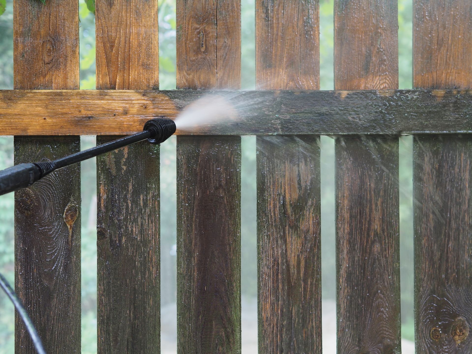 A pressure washer nozzle sprays a stream of water onto a dark, weathered wooden fence, cleaning a light stripe across it.