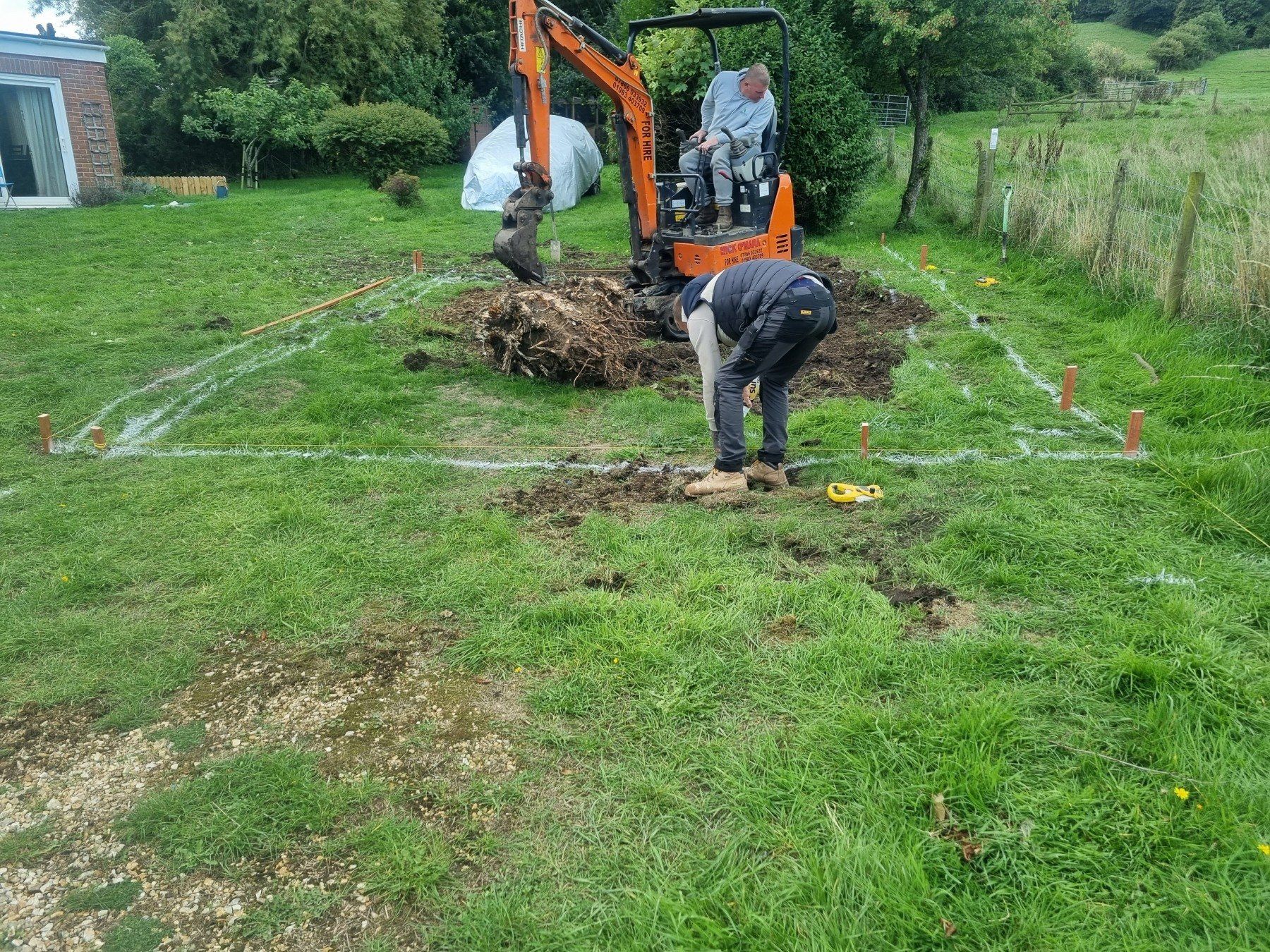 A man is digging a hole in the grass in front of an excavator.