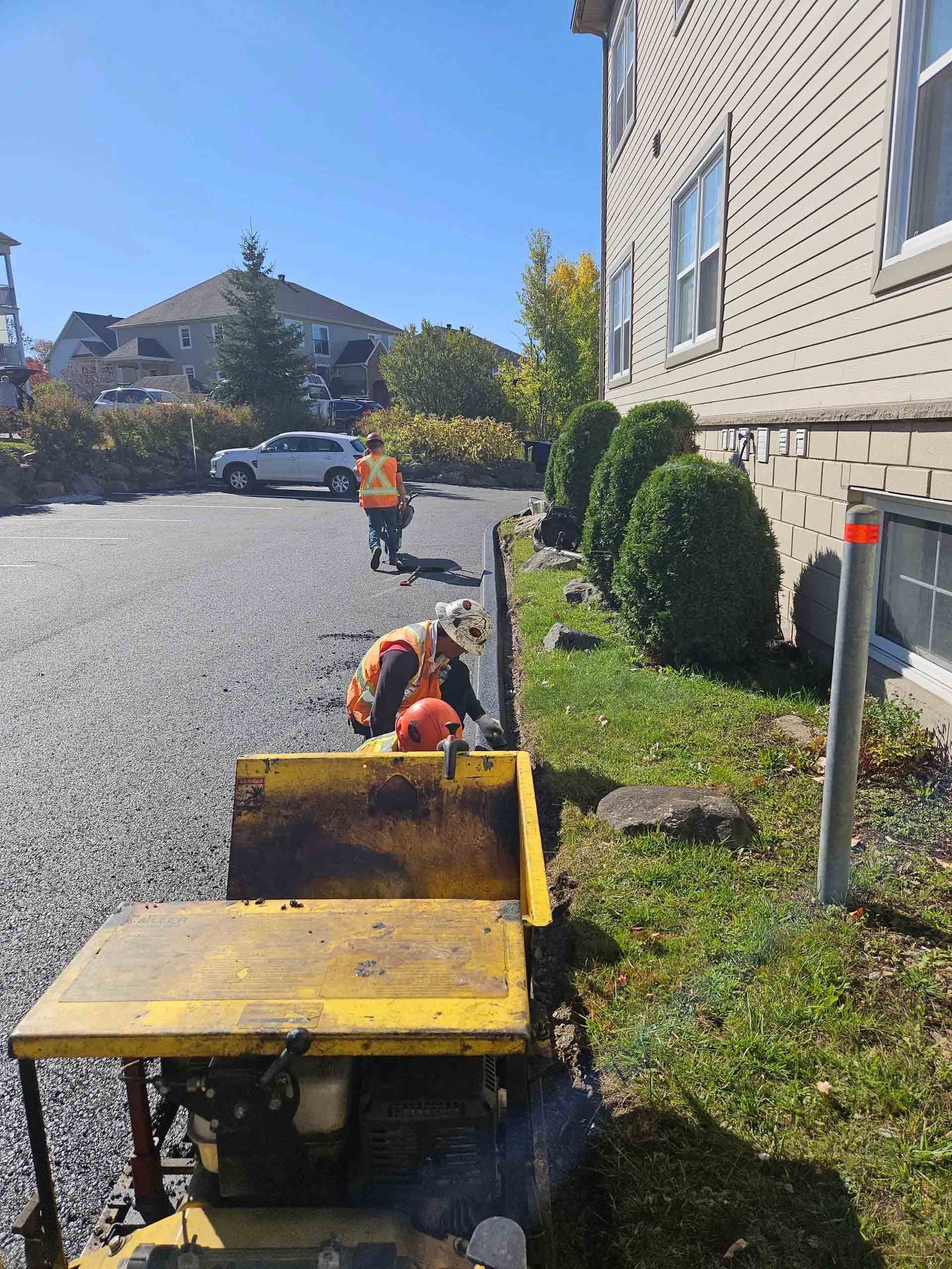 Un ouvrier manœuvre un wagonnet jaune sur du gravier à côté d'un bâtiment, avec un autre ouvrier en gilet orange devant lui.