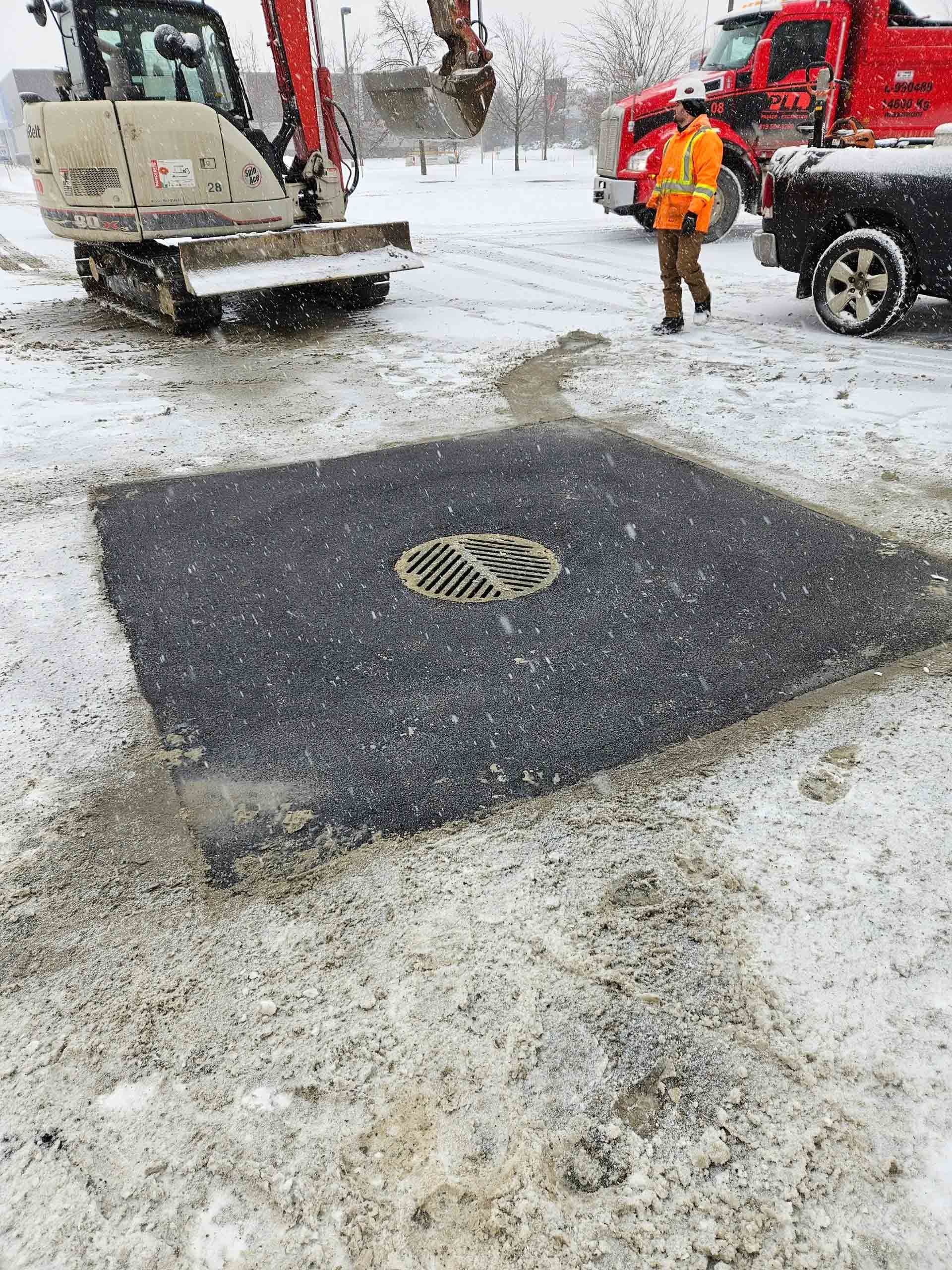 Scène de travaux routiers sous la neige : un ouvrier se trouve près d’un camion et d’une plaque d’égout fraîchement réparée.