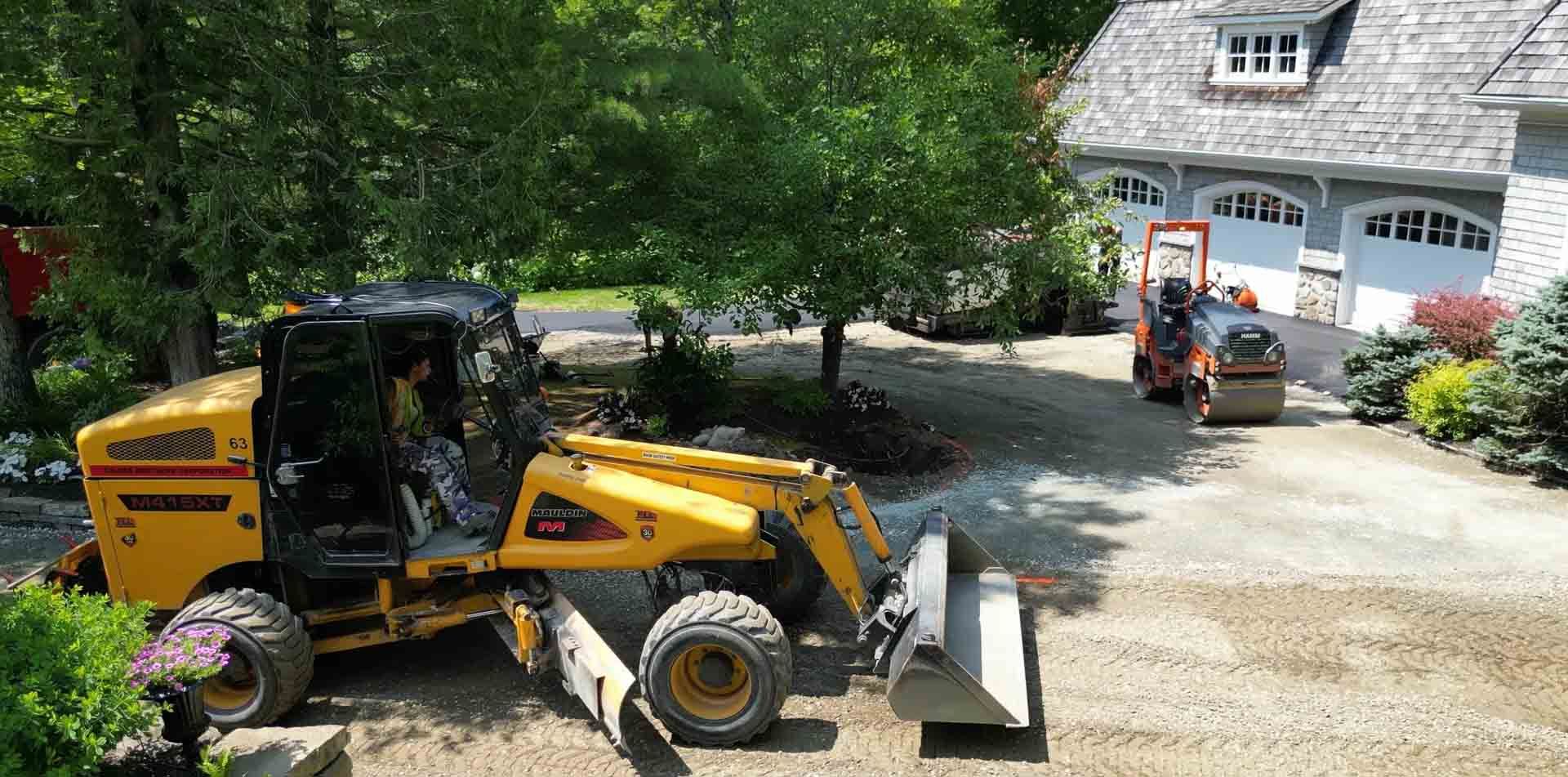 Une chargeuse compacte jaune est garée sur une allée de gravier à côté d'une maison, avec une excavatrice orange en arrière-plan.