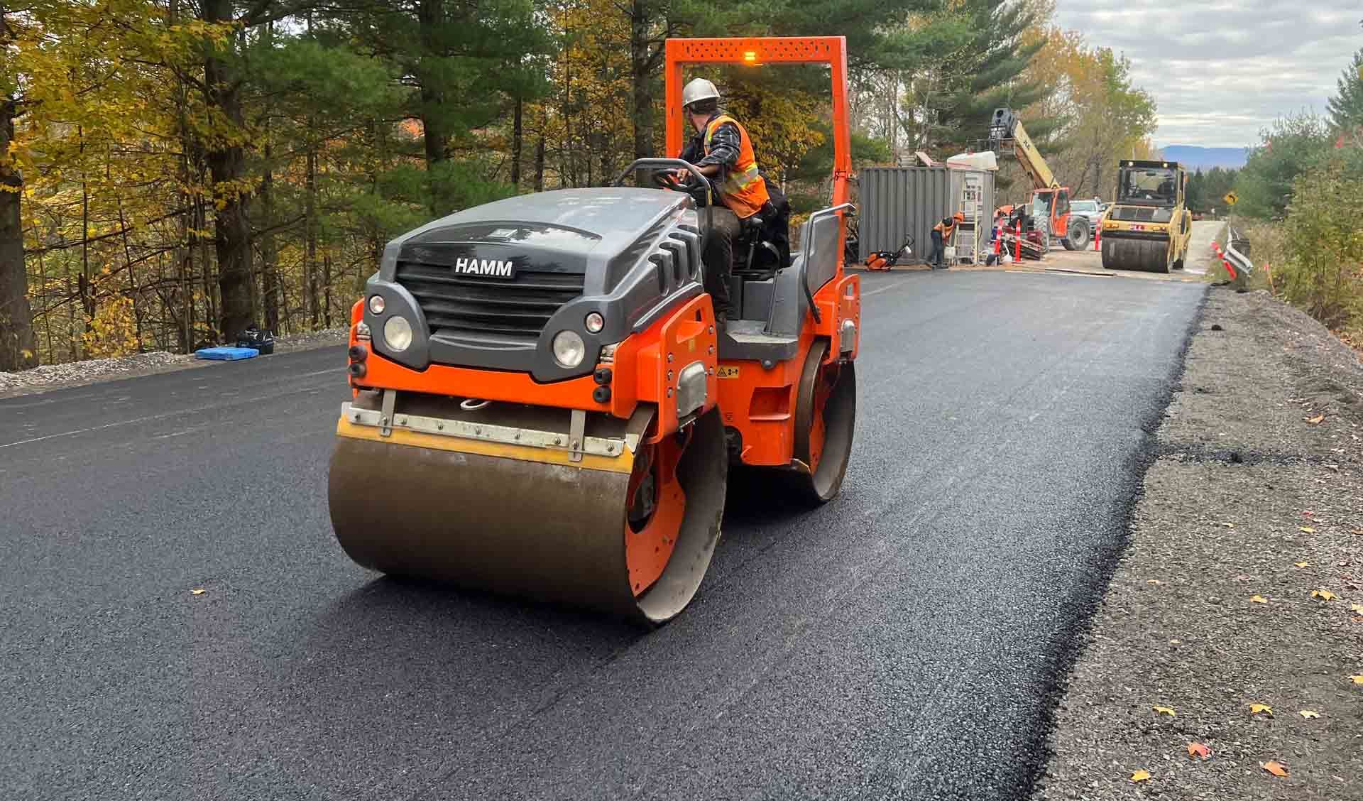 Un rouleau compresseur orange asphalte une route mouillée, avec des ouvriers et des engins de chantier derrière.