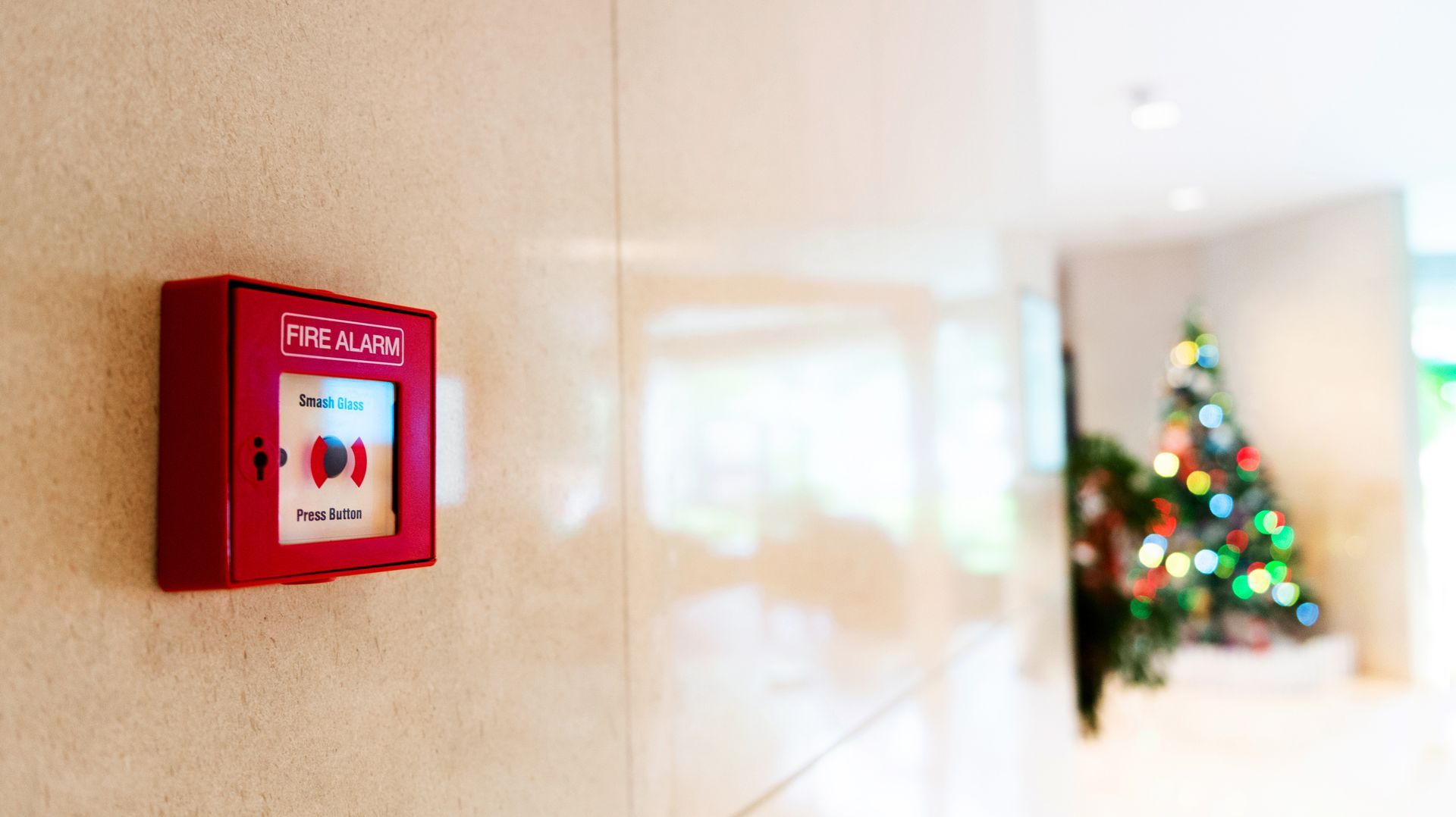 Red fire alarm on beige wall, hallway with decorated Christmas tree in background. Red fire alarm on beige wall, hallway with decorated Christmas tree in background.