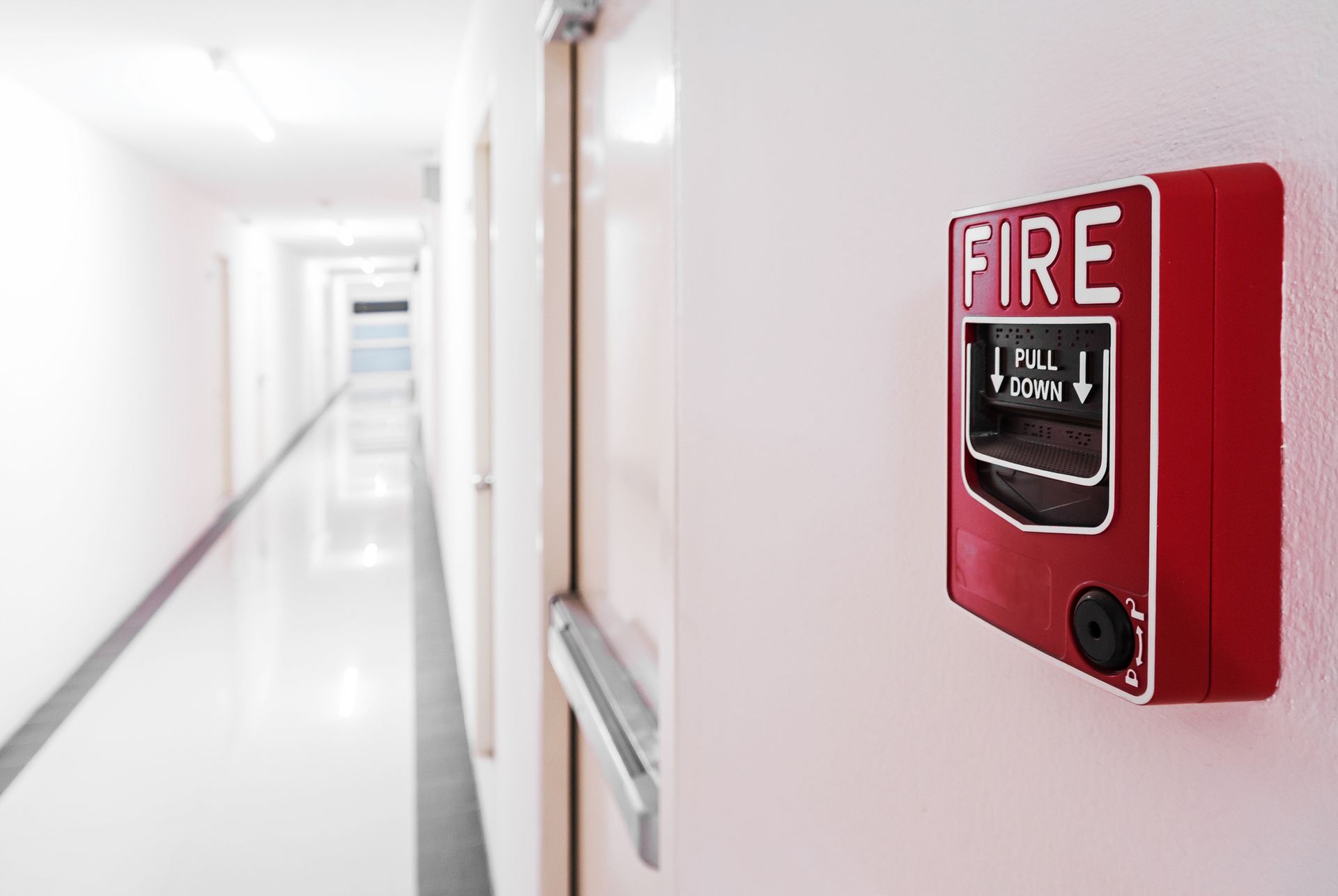 Red fire alarm on a white wall in a bright, long hallway.