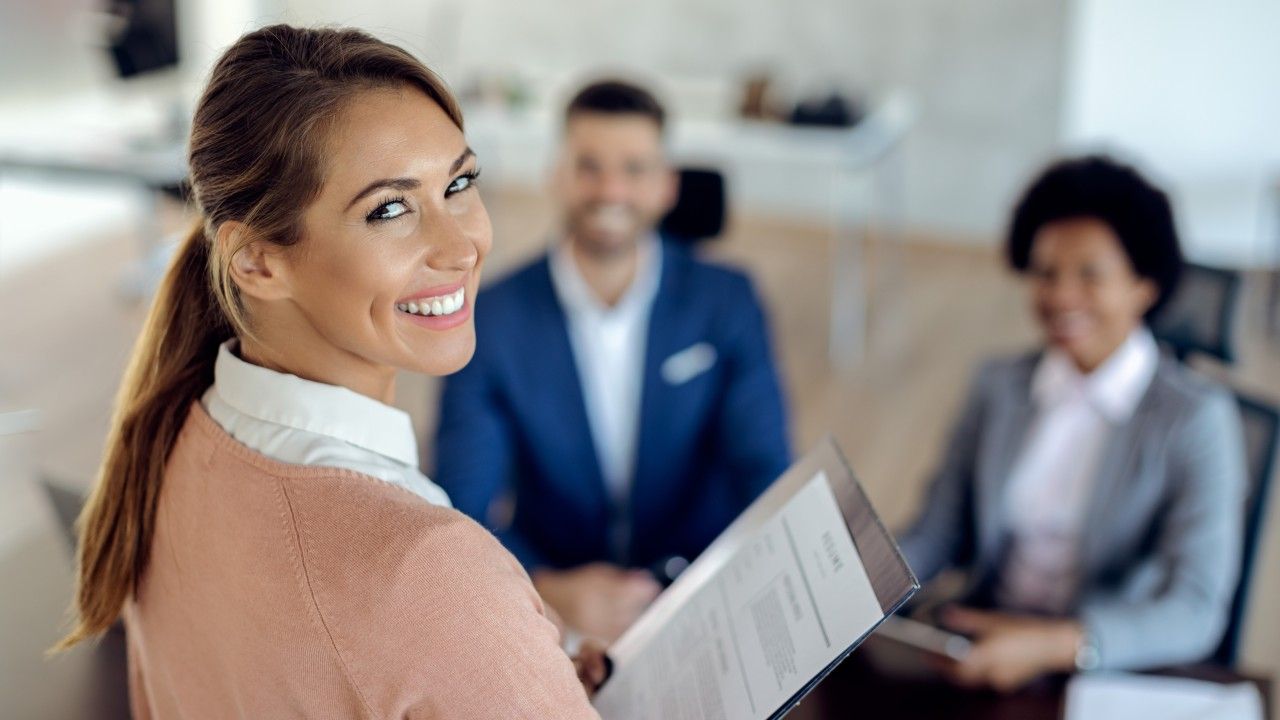 Woman smiles at the camera, holding a document, during a job interview with two others in an office.