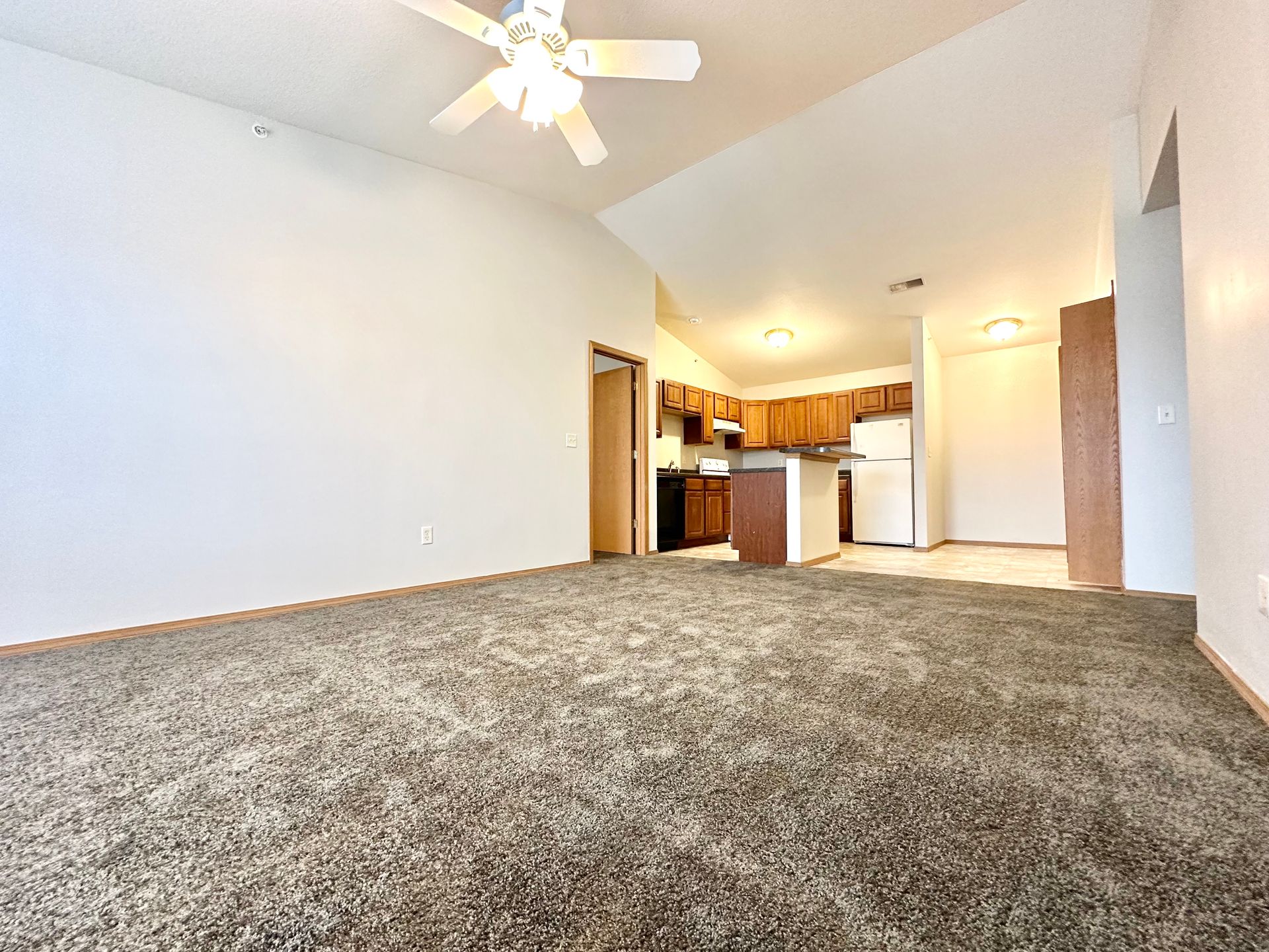 an empty living room with a ceiling fan and a kitchen in the background .