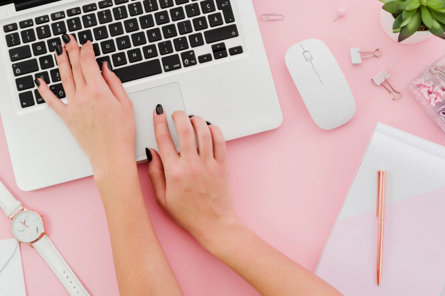 Hands typing on a laptop with a white mouse, watch, and office supplies on a pink surface.