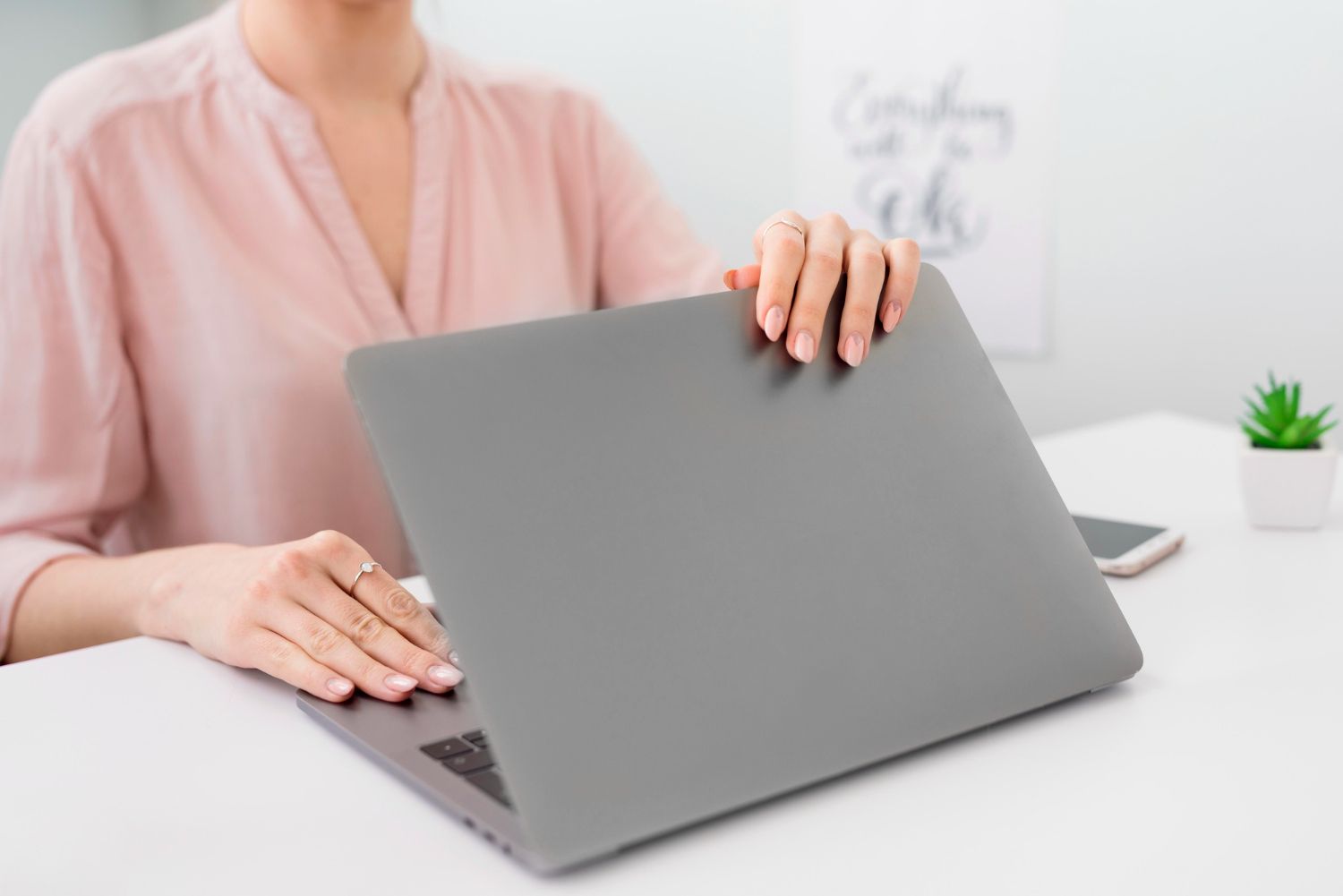 Woman opening a laptop on a white desk, wearing a pink shirt, in a bright room.