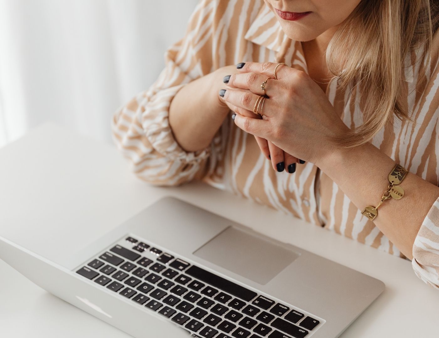 Woman at a laptop, hands on desk. She wears a striped shirt, rings and a bracelet. Black nail polish.