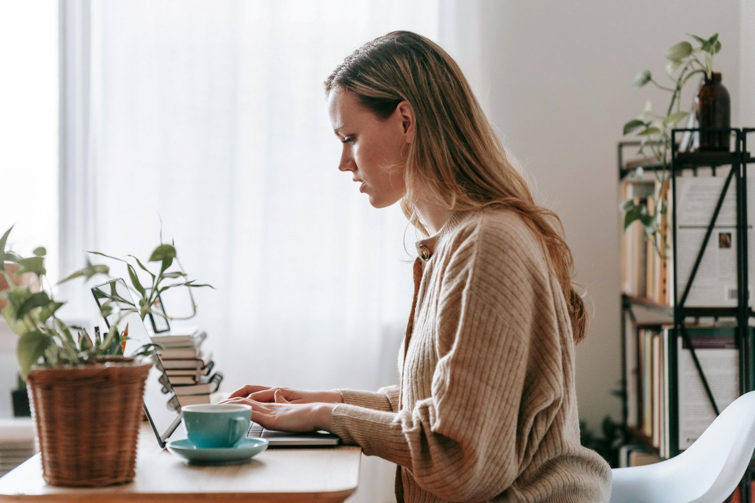 Woman working on laptop at desk, window in background, cup of tea, plants.