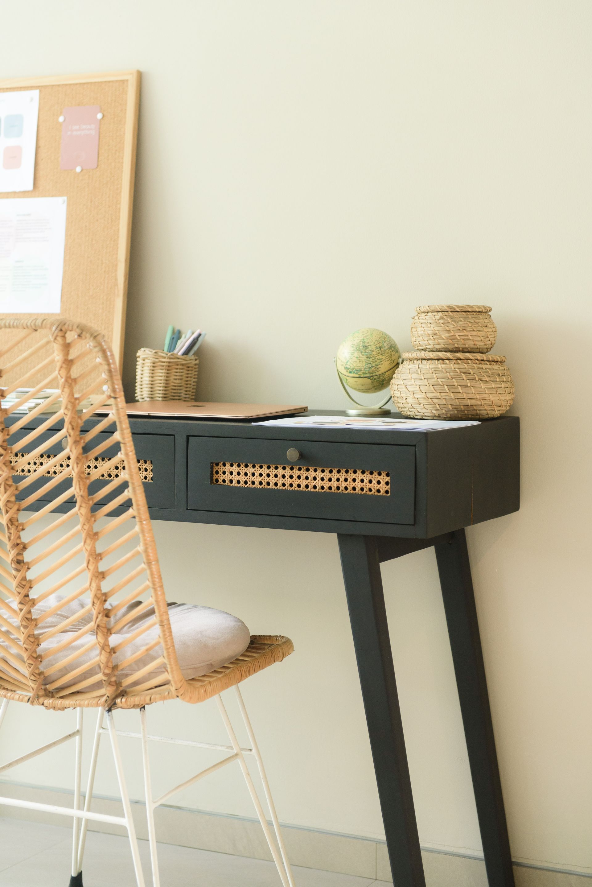 A rattan chair sits at a dark desk with wicker drawer front and corkboard, against a pale wall.