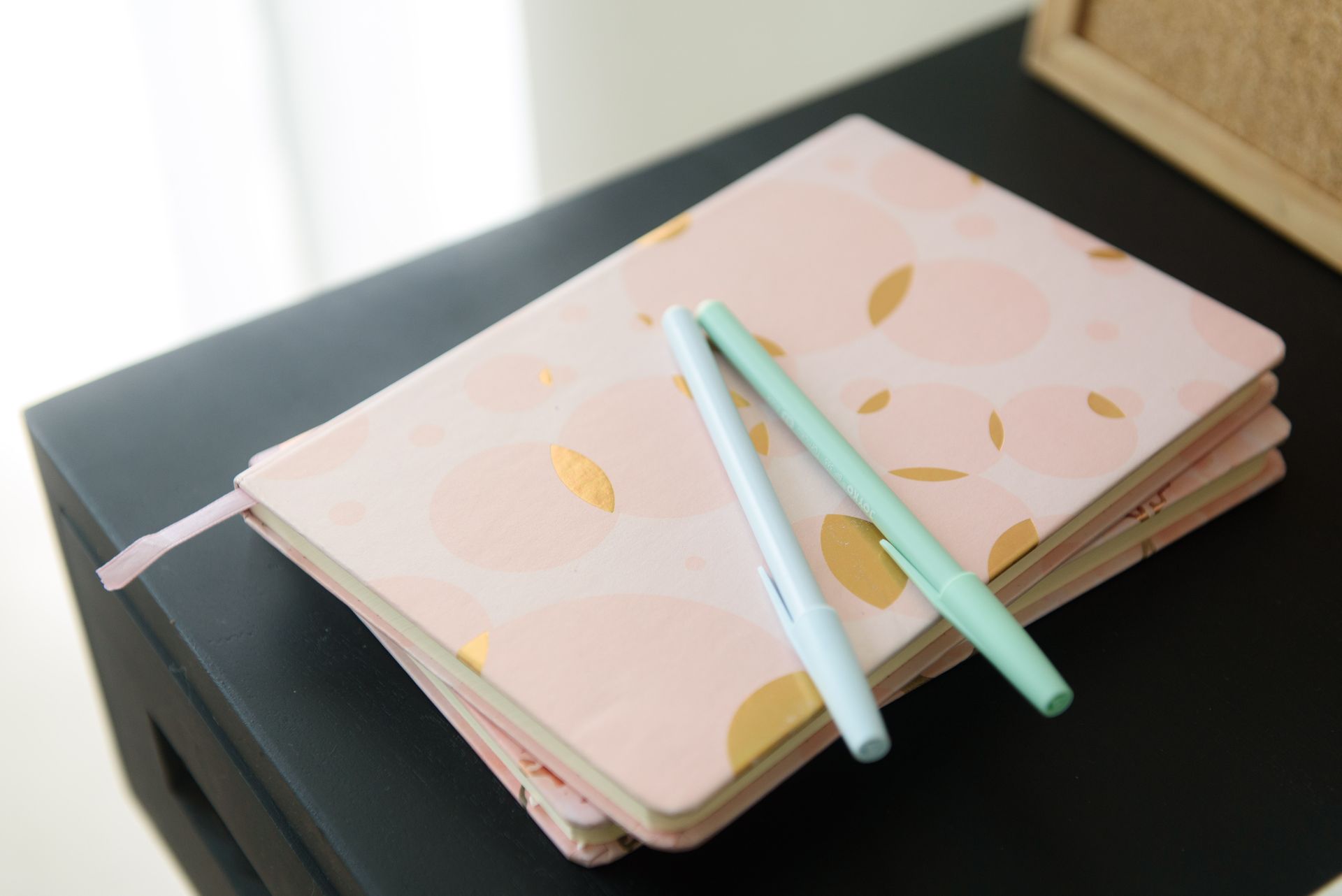 Pink journal with gold circles and two pens on a black table.