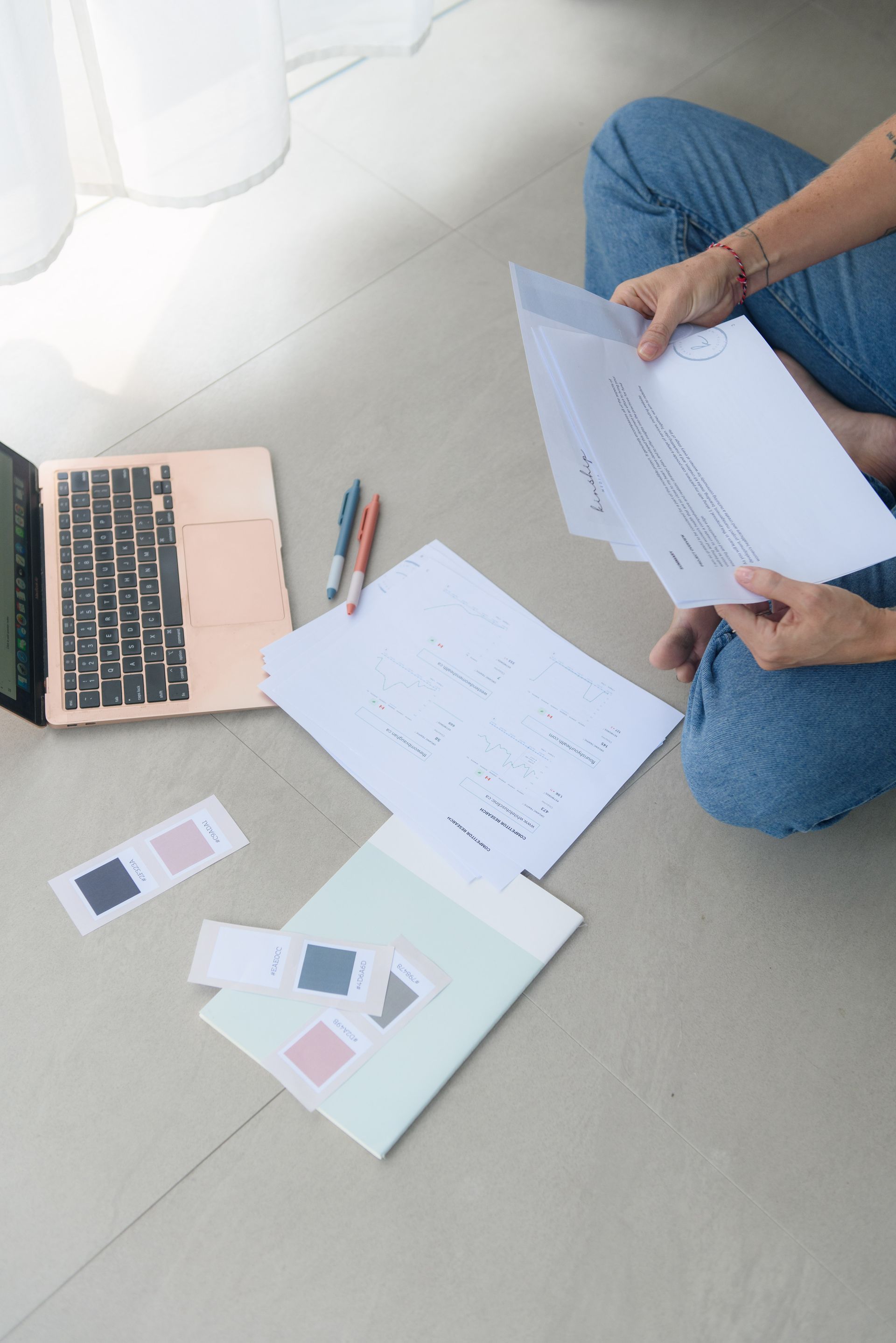 Person sitting on floor with papers, laptop, color swatches, and pens.