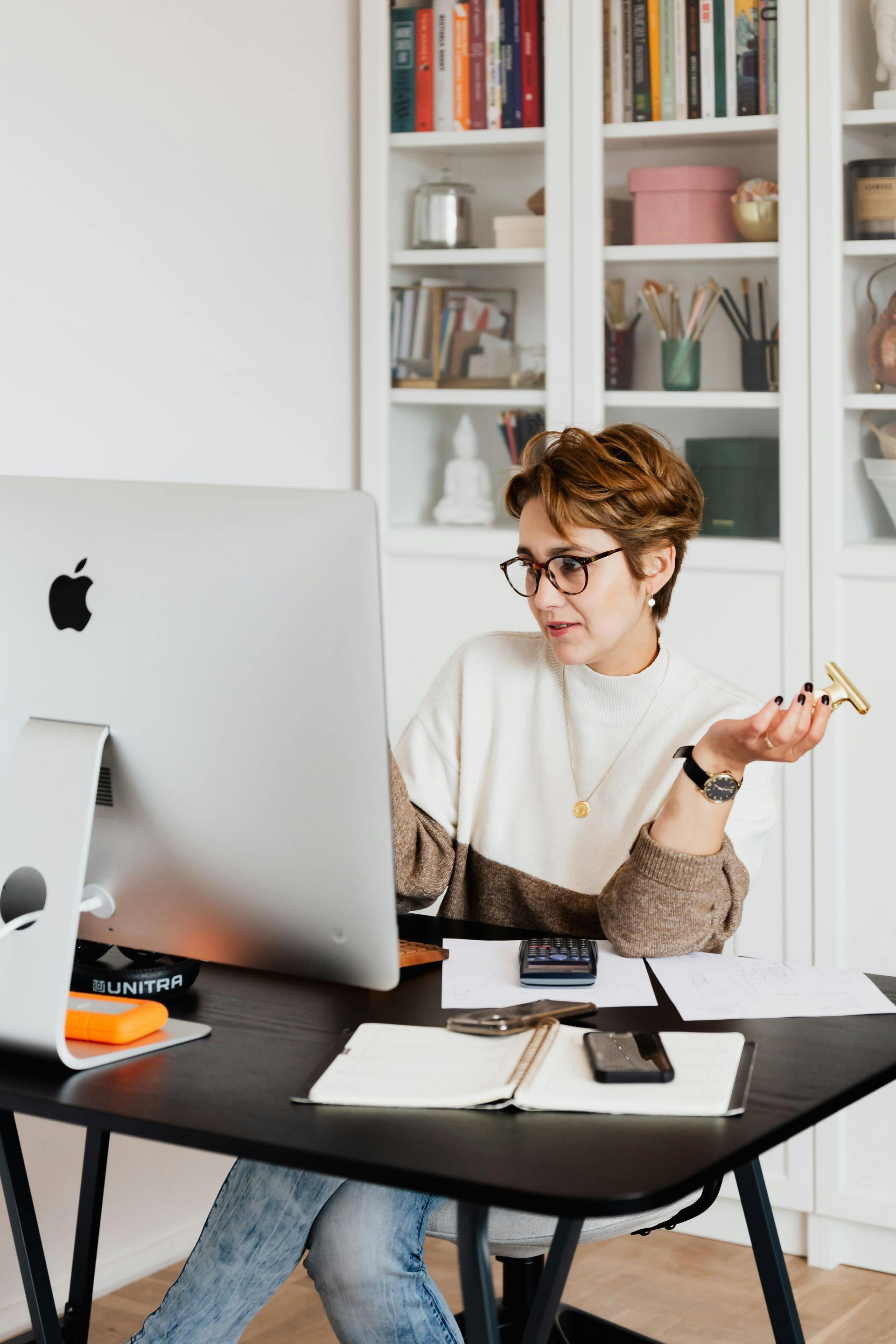 Woman in glasses gestures at a computer screen, seated at a desk. A bookshelf is in the background.