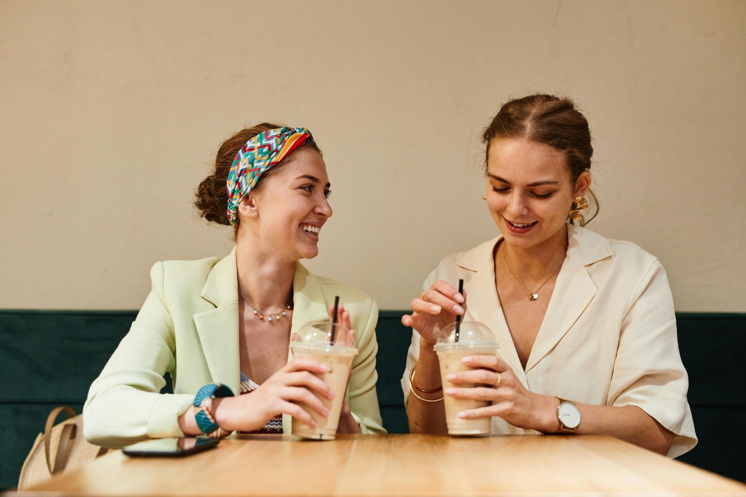 Two women at a cafe, smiling and drinking iced coffee, sitting at a wooden table.
