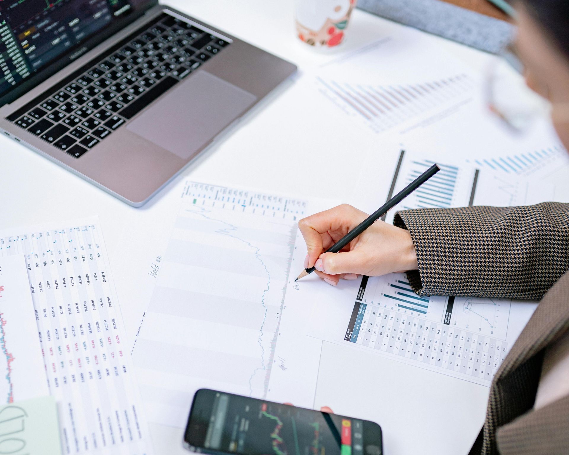 Person writing on paper, laptop, phone with financial data on desk.