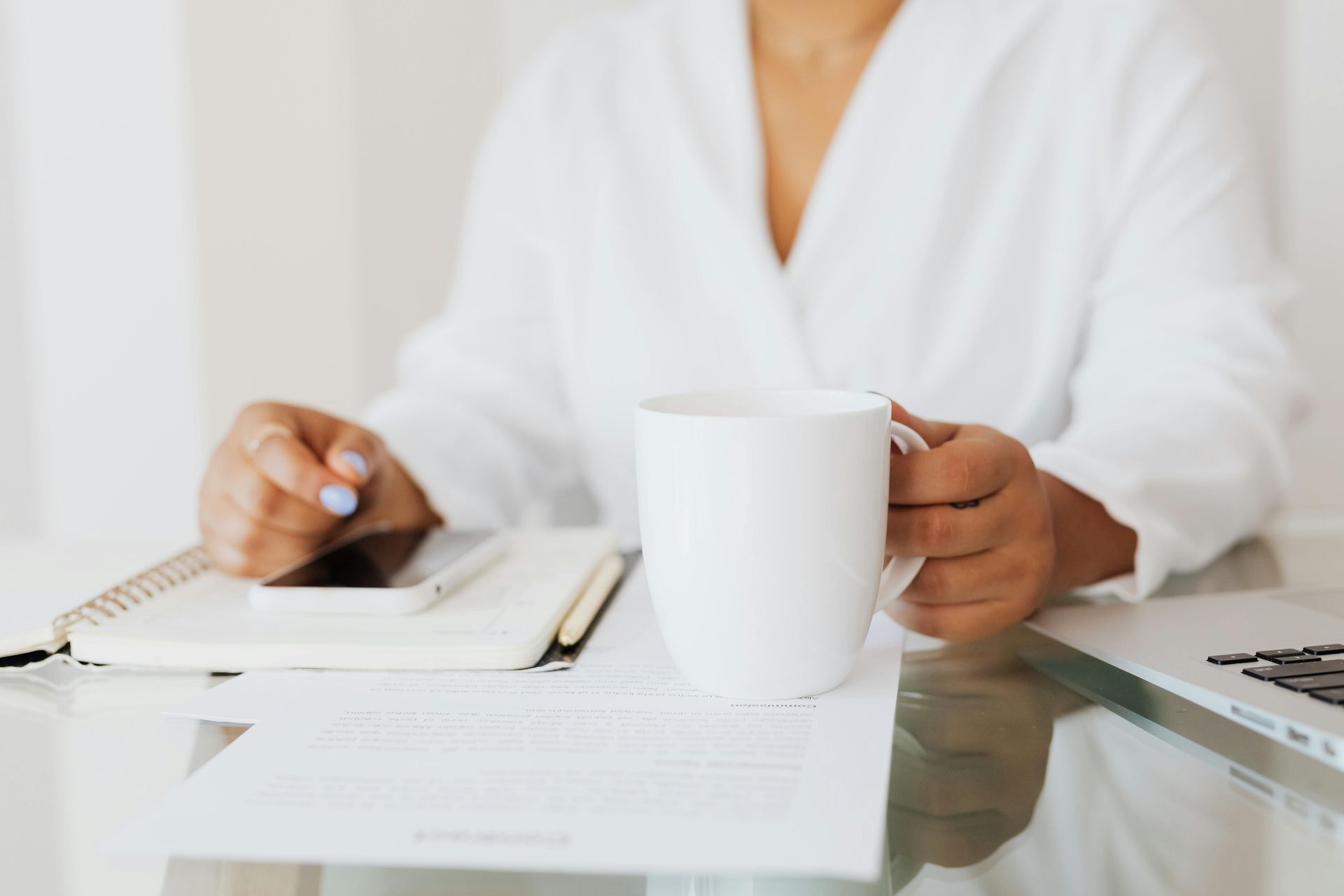 Person in white robe holding white mug at a desk with documents, notebook, phone, and laptop.
