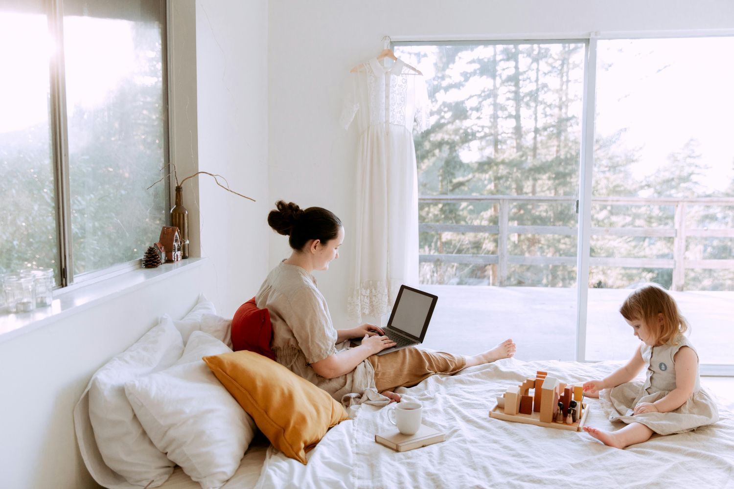 Woman working on laptop in bedroom while child plays with wooden toys. Bright room, large window.