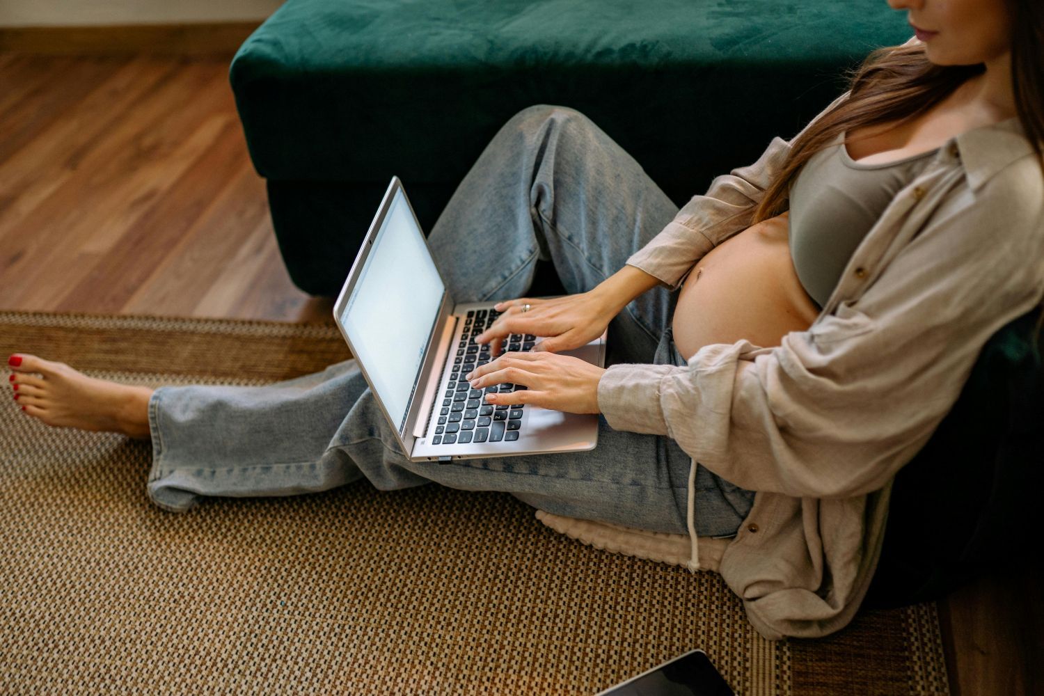 Pregnant person using a laptop while sitting on the floor by a green couch.