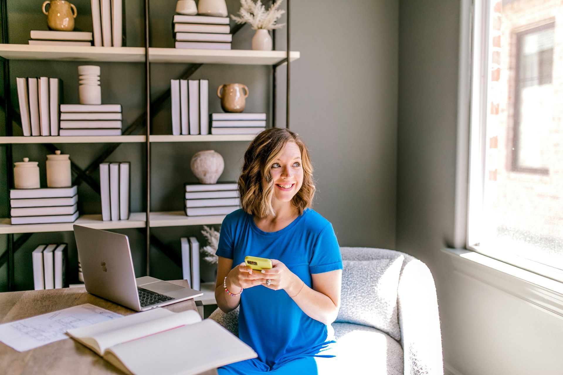 Woman holding a mug beside a laptop in a bright home office with shelves and a large window