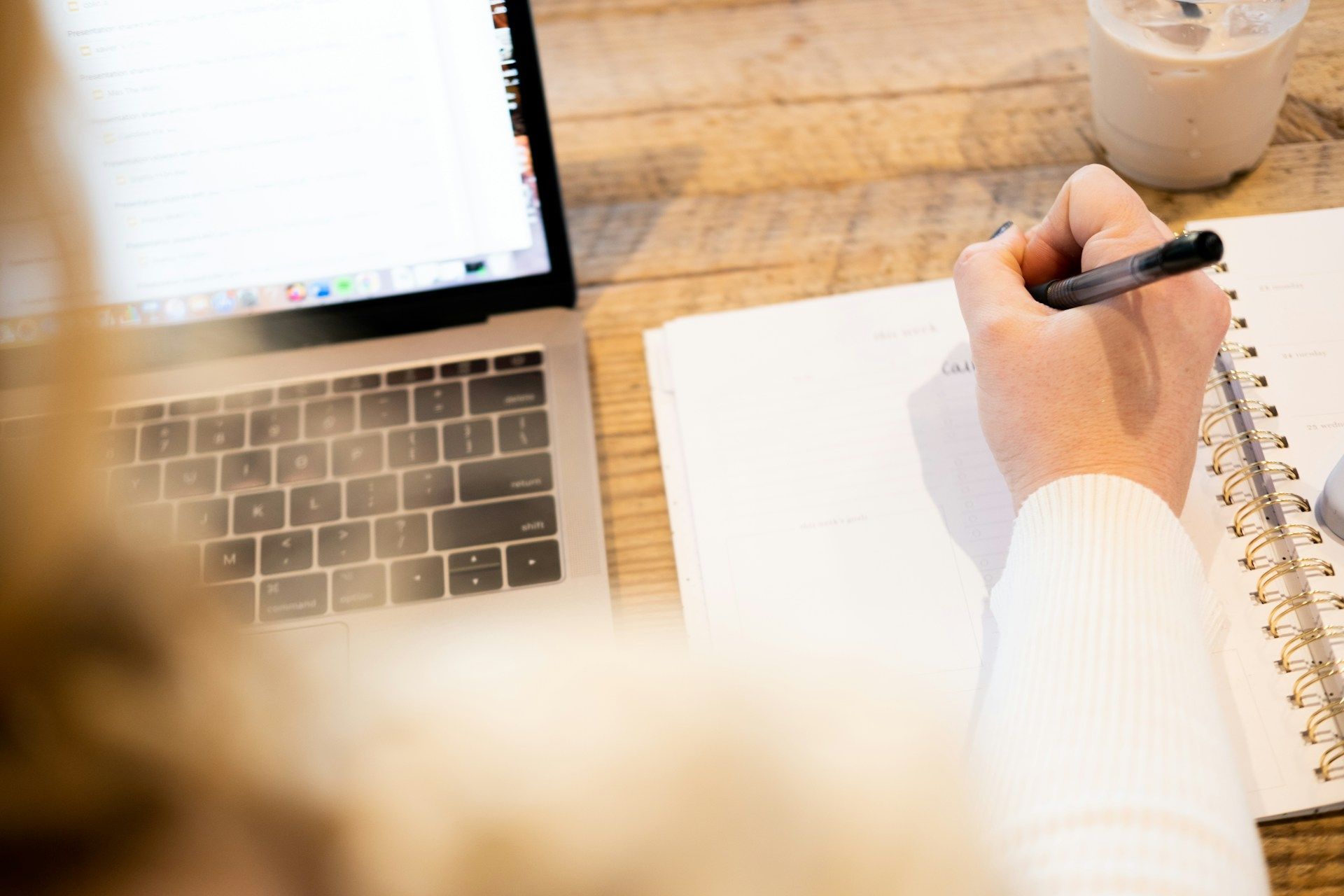 Person writing in a notebook next to a laptop on a wooden table.