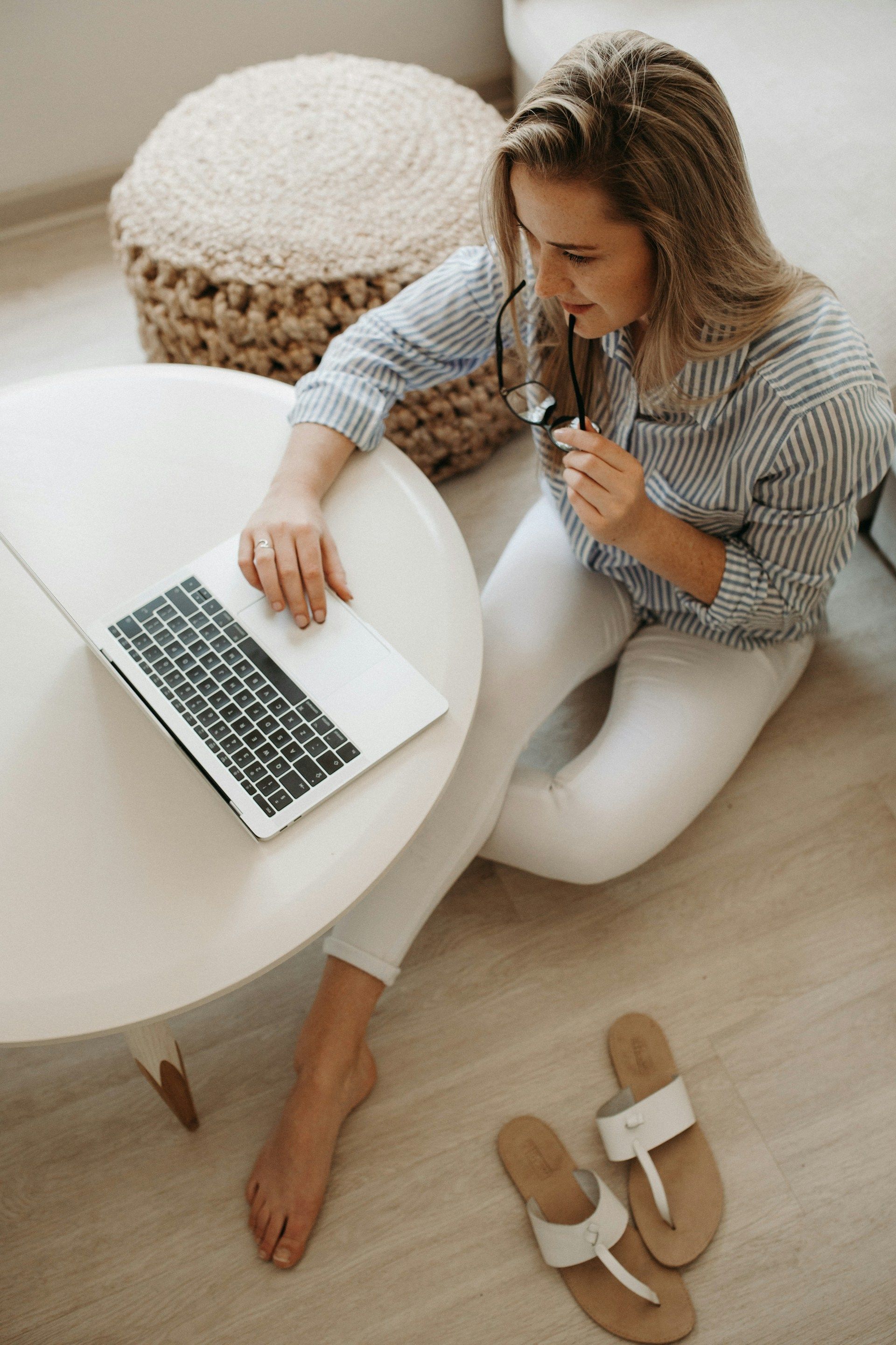 Woman sitting on floor with laptop, holding glasses, near sandals, and round table.