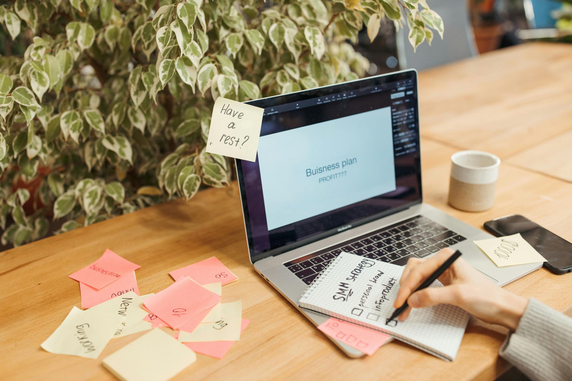 Laptop on wooden desk with sticky notes, hand writing, and plant in the background.